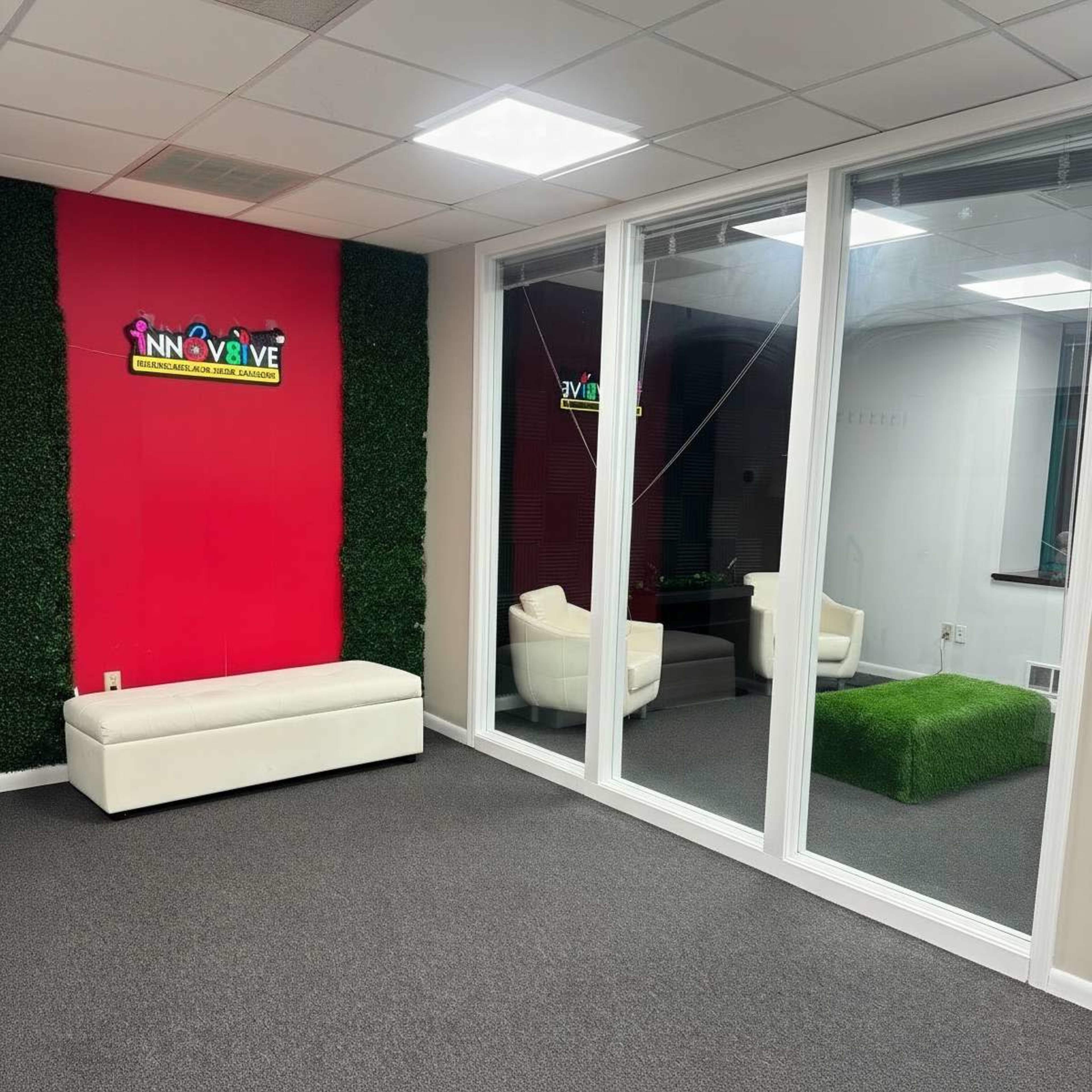 The image shows a modern office space featuring a red accent wall with a logo, white seating, and glass walls separating an area with green decor.