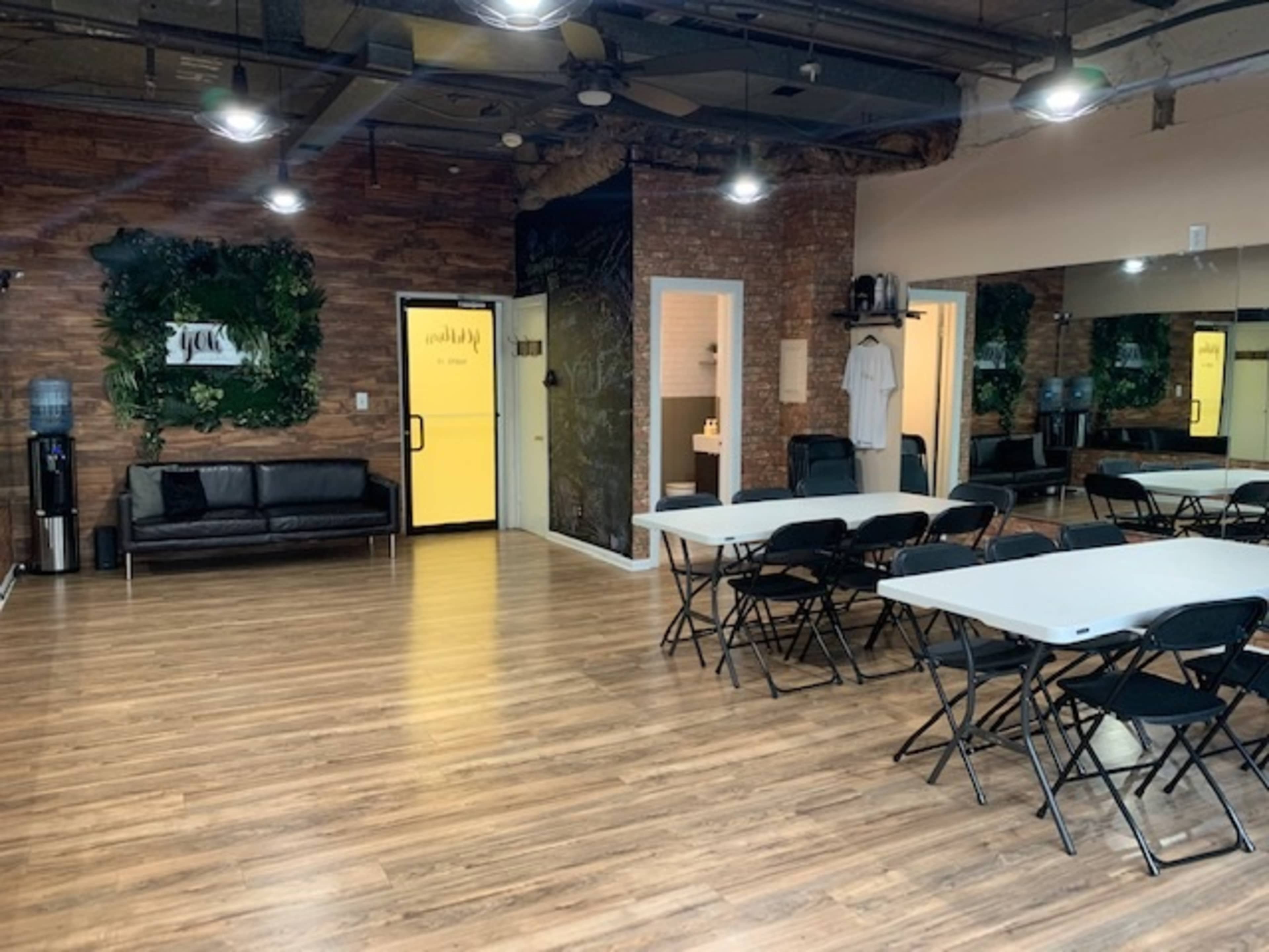 The image shows a spacious room with wooden flooring, a black couch, several folding tables, and a mirrored wall, illuminated by ceiling lights.