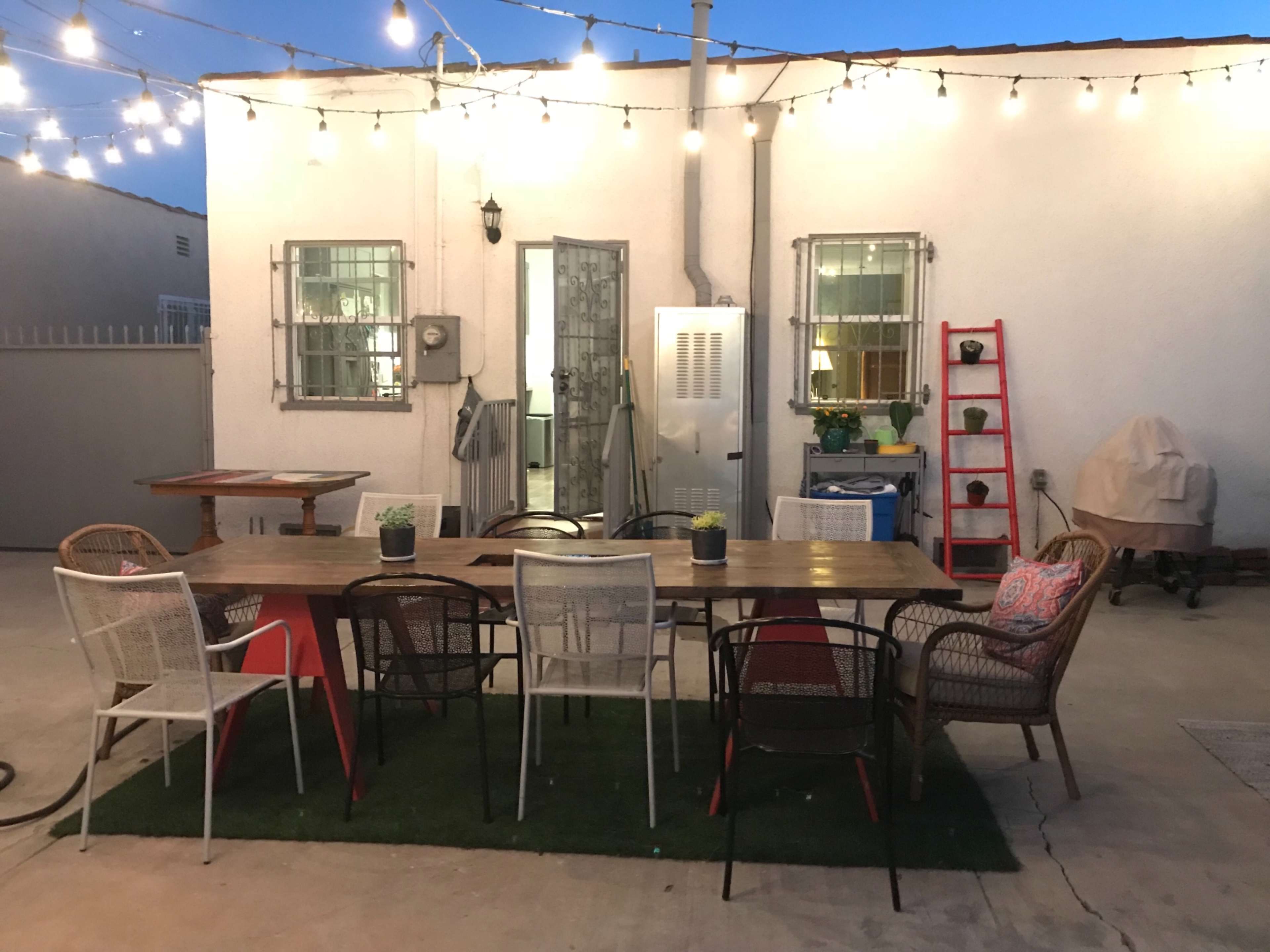 A dining table surrounded by chairs is set in a backyard with string lights overhead, leading to a white building with multiple windows.