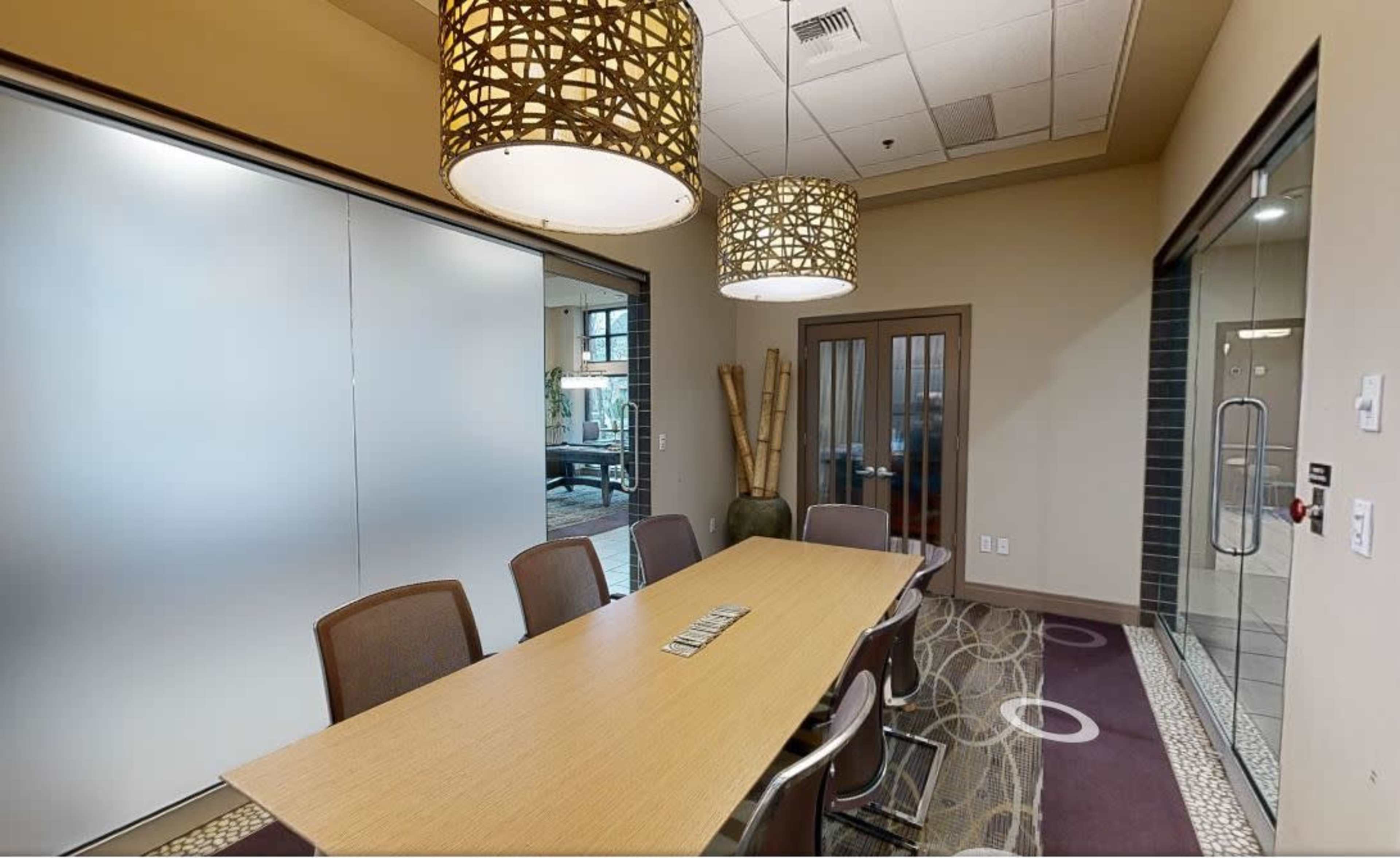 A conference room features a long wooden table surrounded by chairs, with two decorative pendant lights hanging above and glass-paneled doors leading to another space.