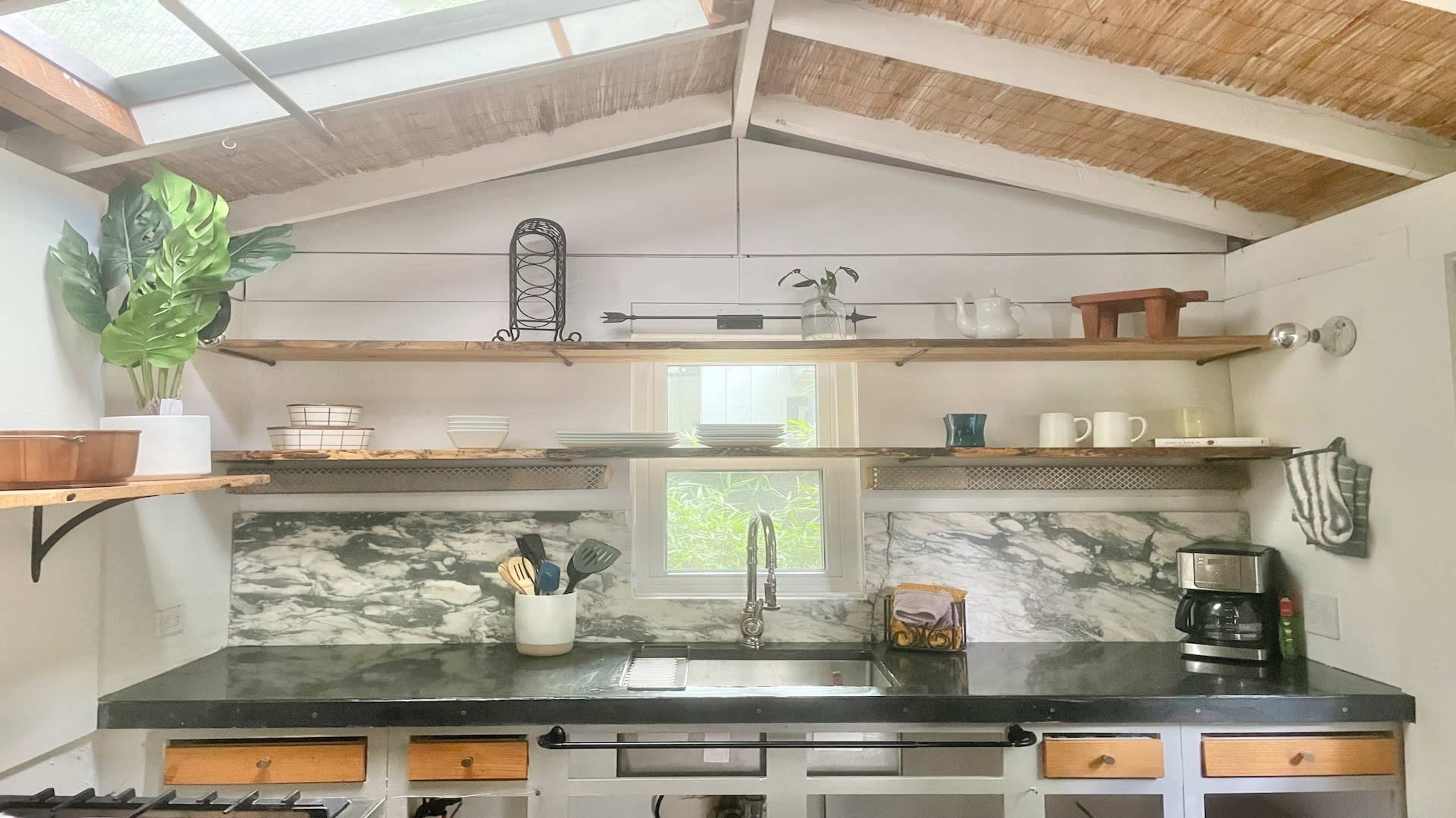 A kitchen with open shelves displaying dishware and plants, a black countertop, and a window allowing natural light to enter.