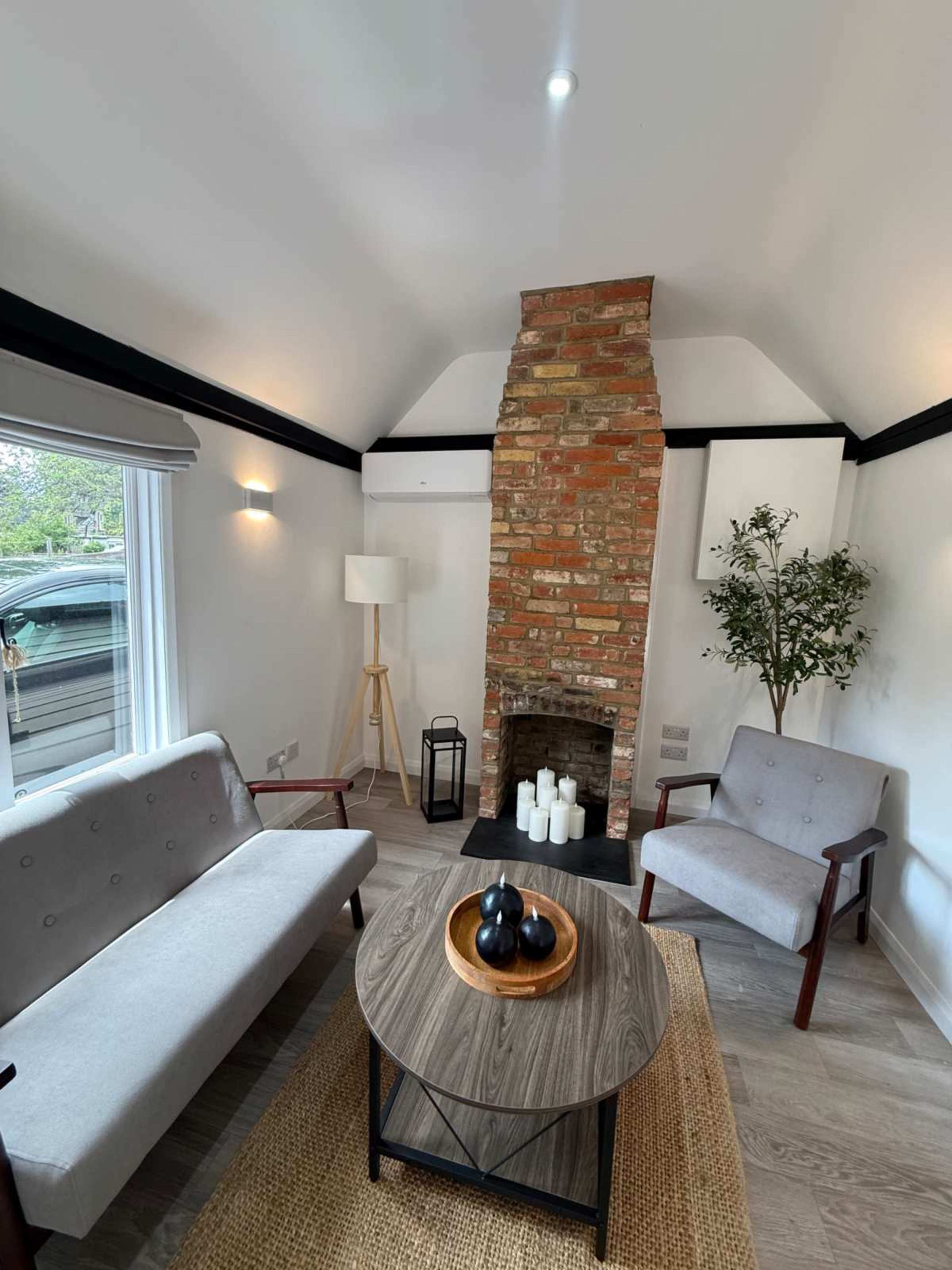 The image shows a modern living room featuring a brick fireplace, two grey armchairs, a round wooden coffee table, and a cozy rug.