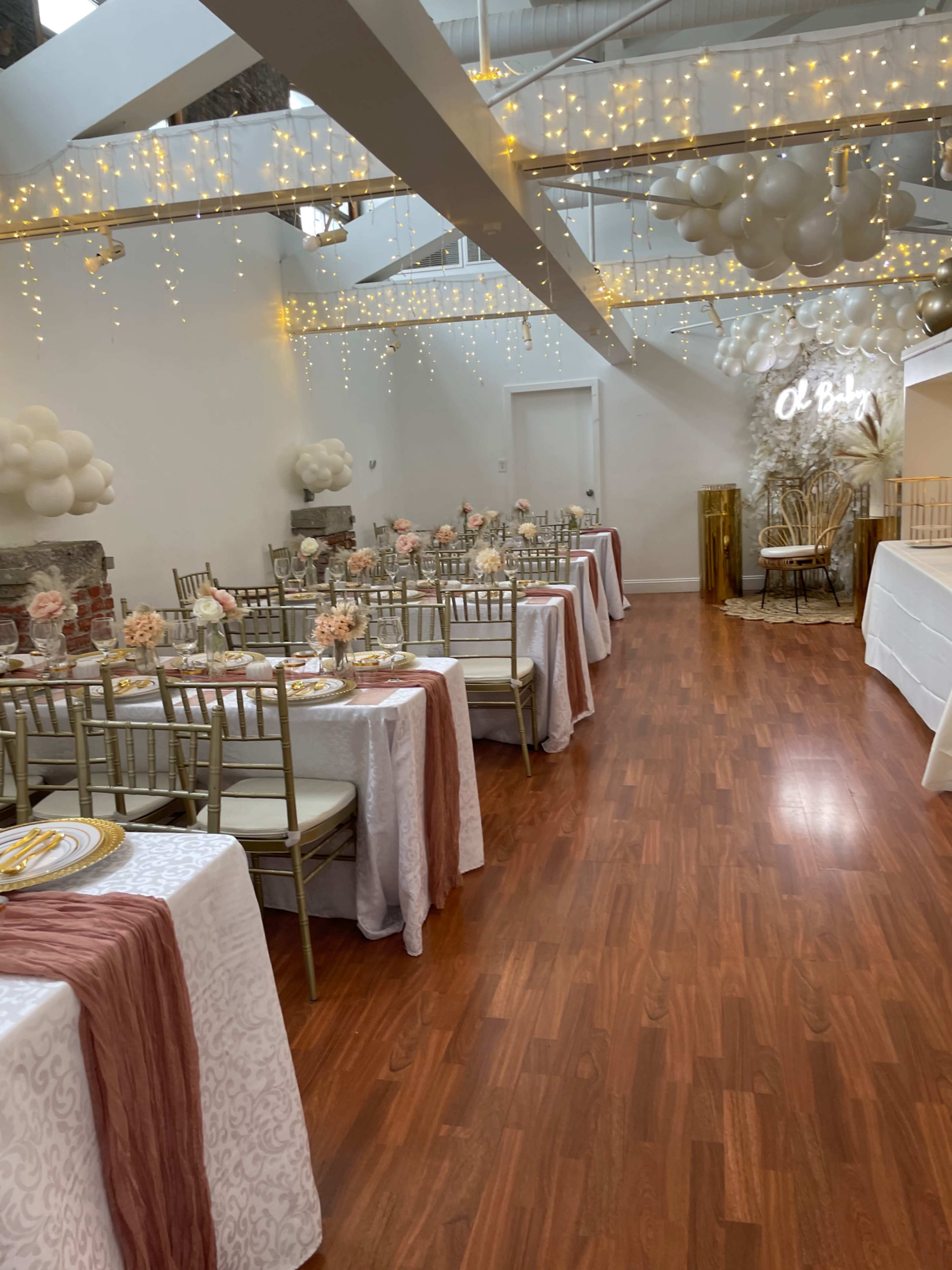 The image shows a decorated event space with tables set for a gathering, featuring floral centerpieces and string lights hanging from the ceiling.
