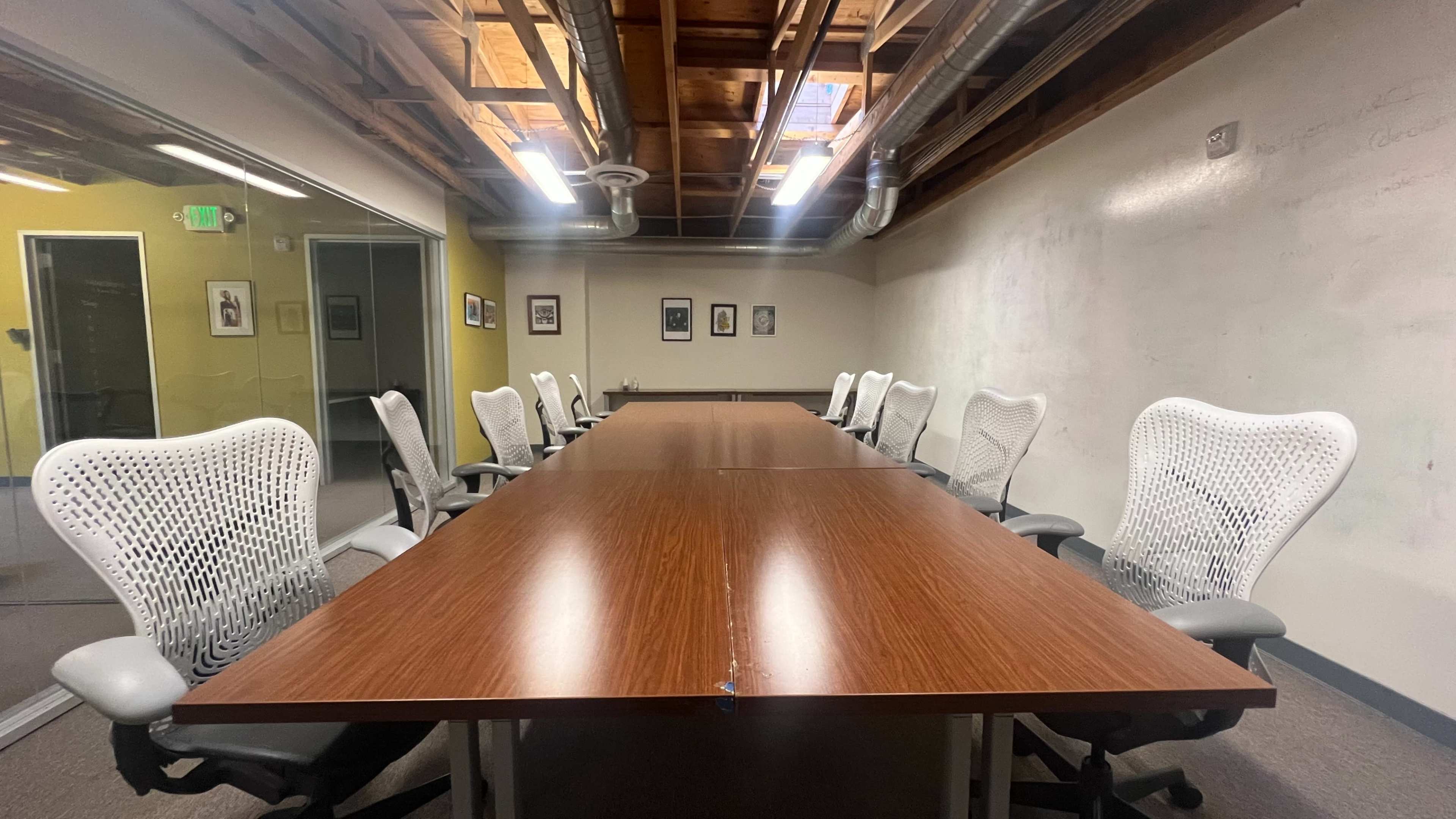 A large conference room features a long wooden table surrounded by white ergonomic chairs, with walls showcasing framed pictures.
