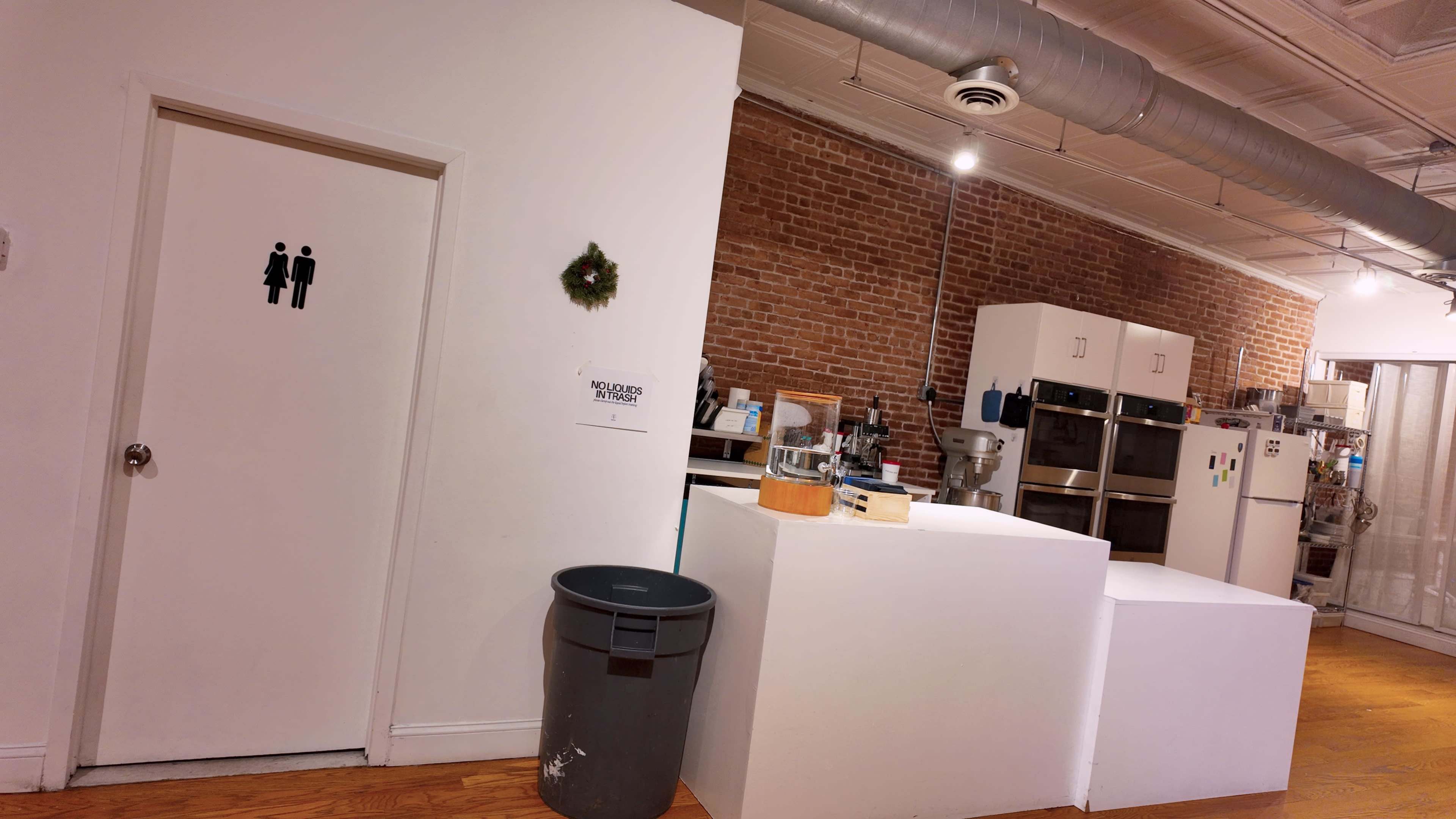 The image shows an interior space with a bathroom door marked with a gender symbol, a kitchen area with appliances, and a trash can beside a white partition.
