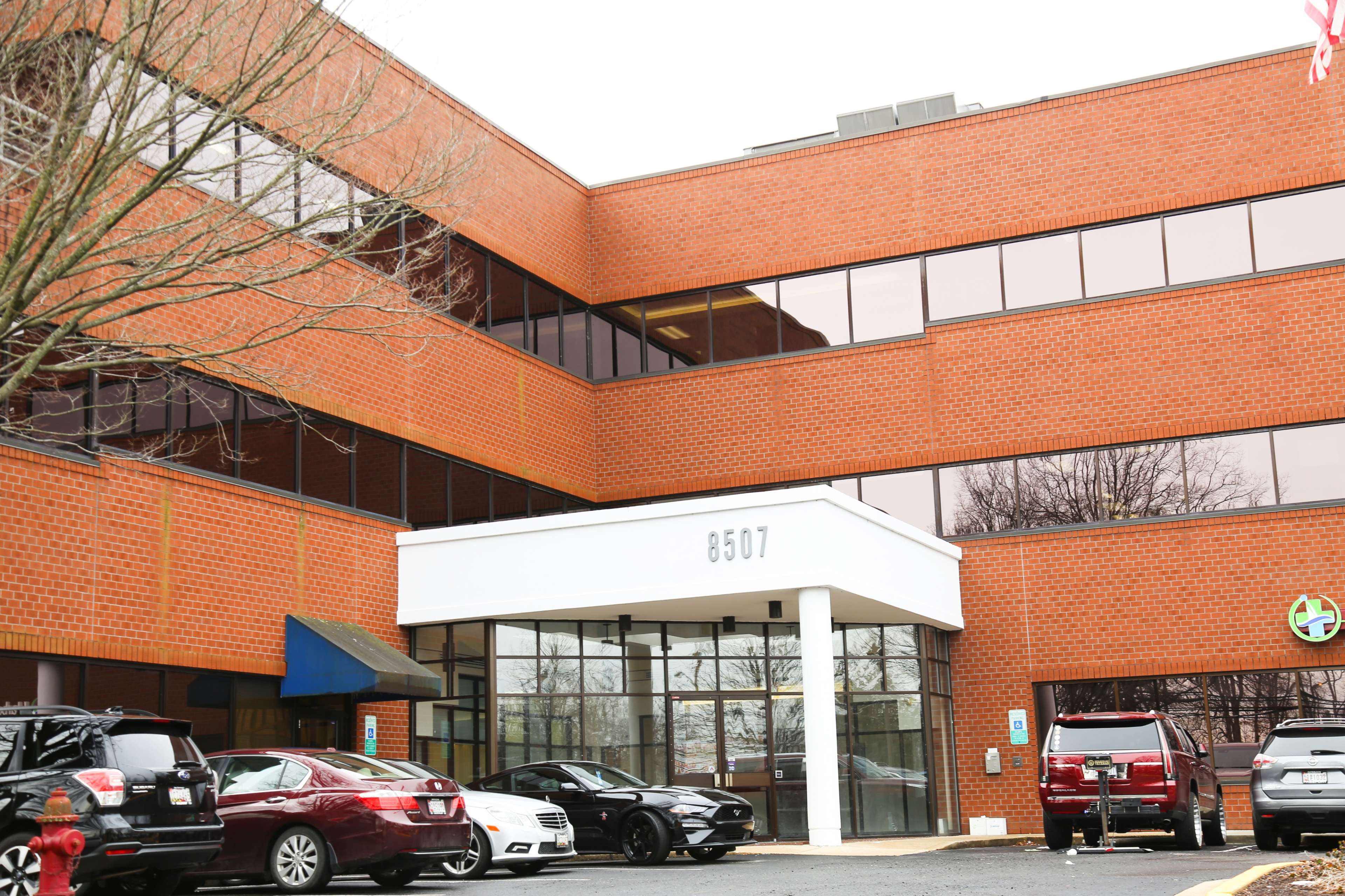 A three-story brick office building with large windows and a covered entrance, flanked by several parked cars in the foreground.