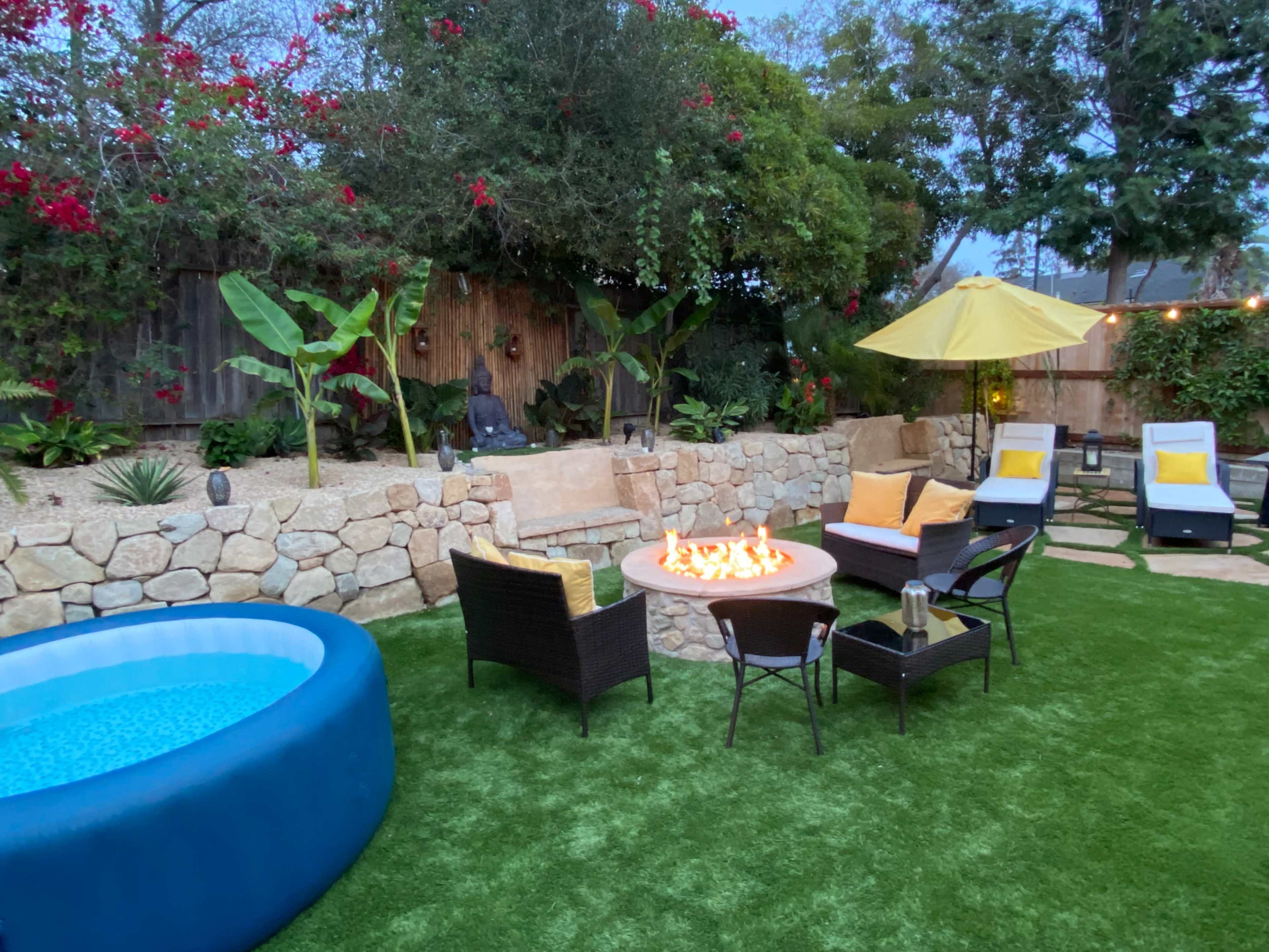 A landscaped backyard features a circular fire pit surrounded by seating, a blue inflatable pool, a yellow umbrella, and various plants.