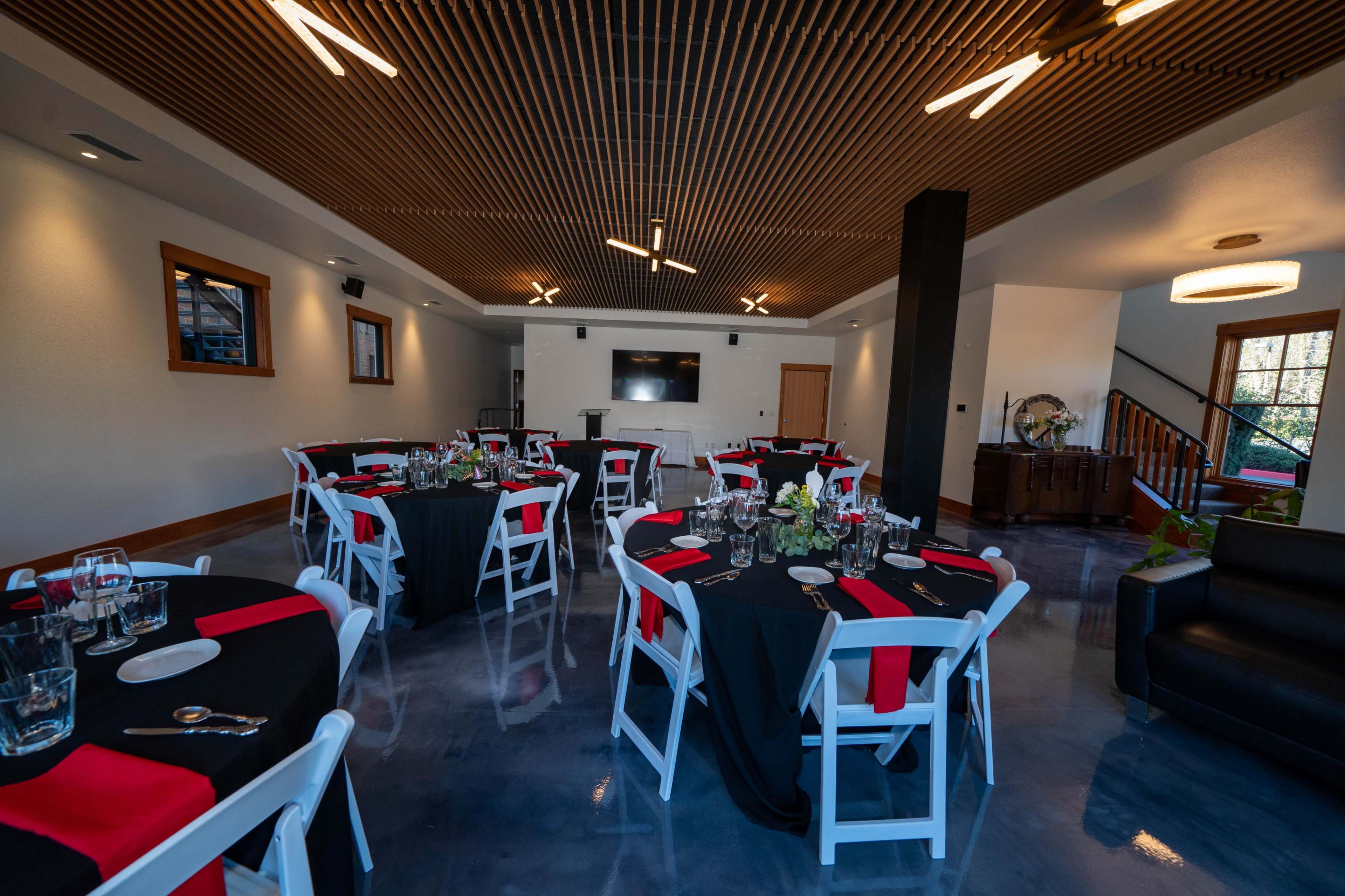 A dining area set with round tables covered in black tablecloths and red accents, arranged around a central space with a flat-screen TV and minimalist decor.