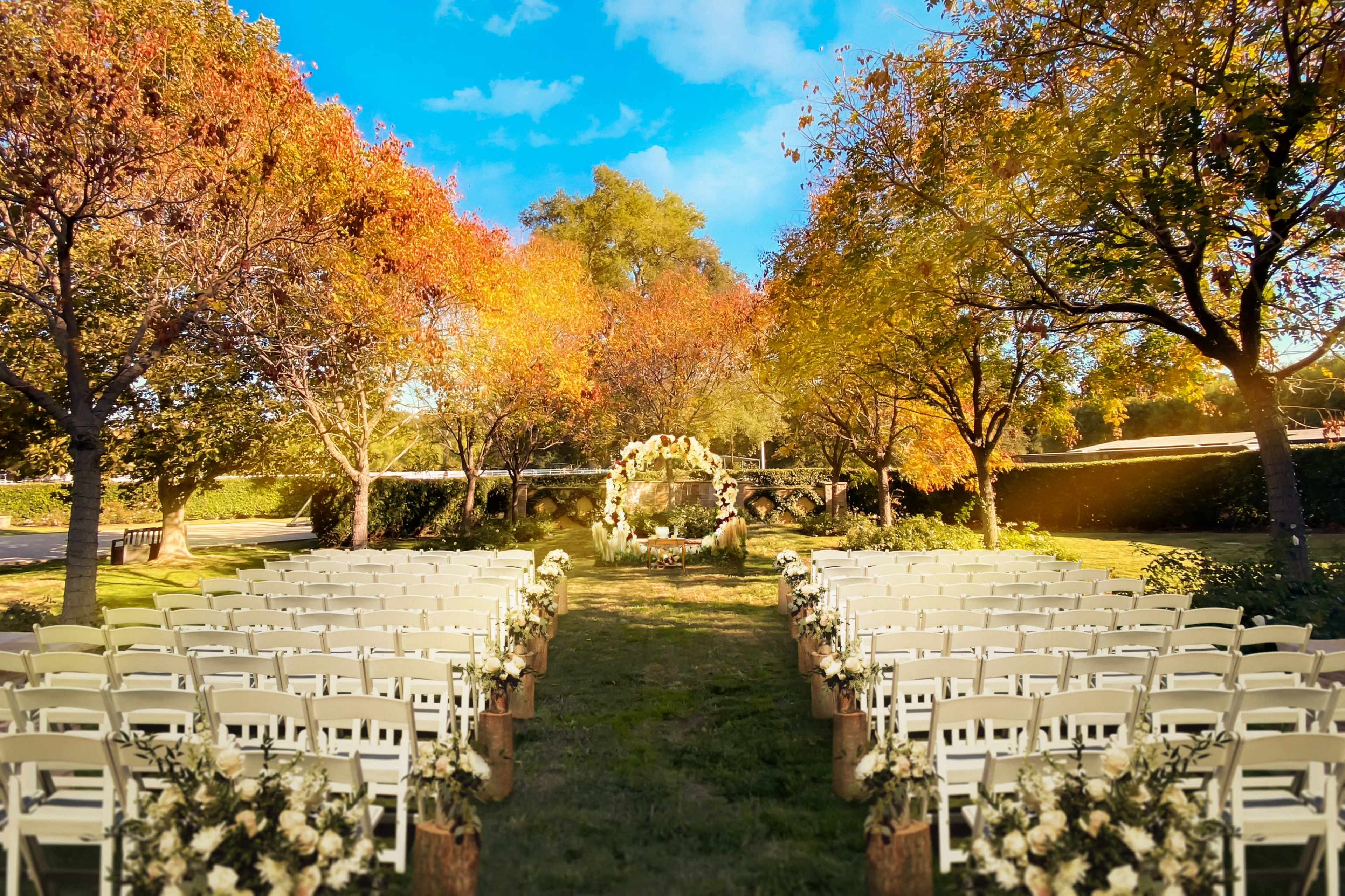 A wedding ceremony setup features rows of white chairs arranged on a grassy area surrounded by trees with autumn foliage.