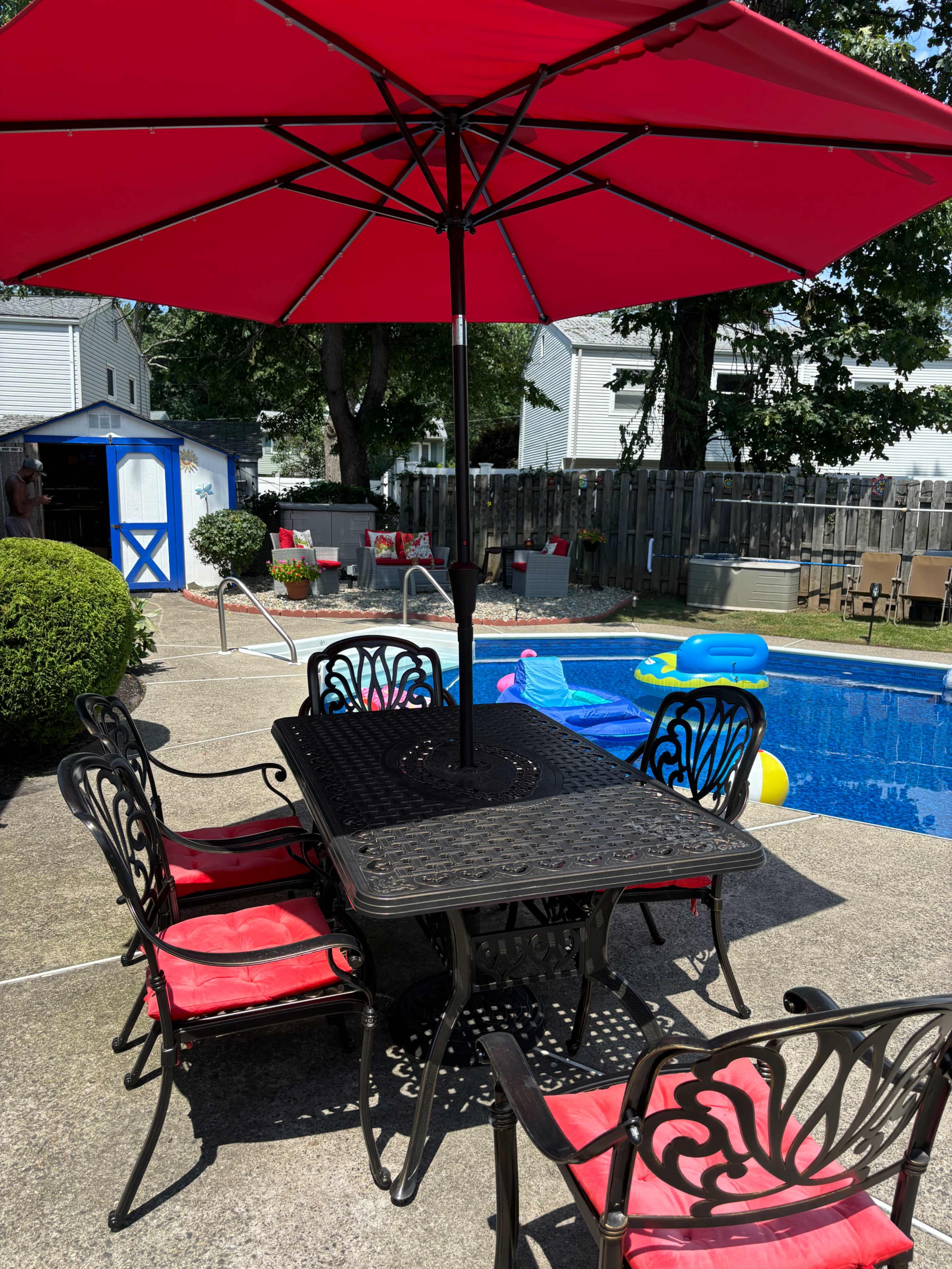 A patio table with red cushions and an umbrella stands next to a swimming pool, surrounded by green bushes and a yard with various items in the background.