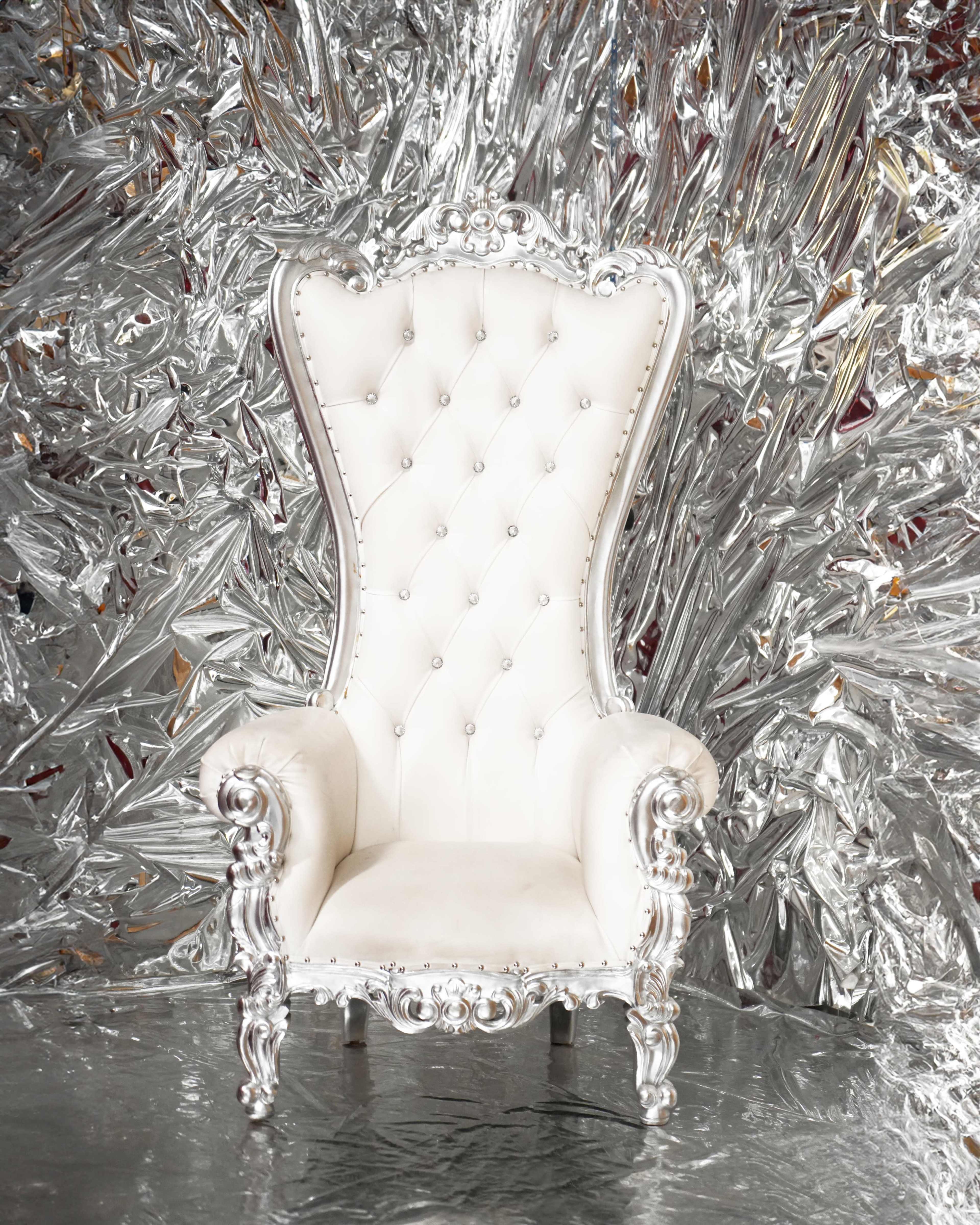 A white, tufted high-back chair with ornate silver legs is placed against a shiny, crinkled metallic backdrop.