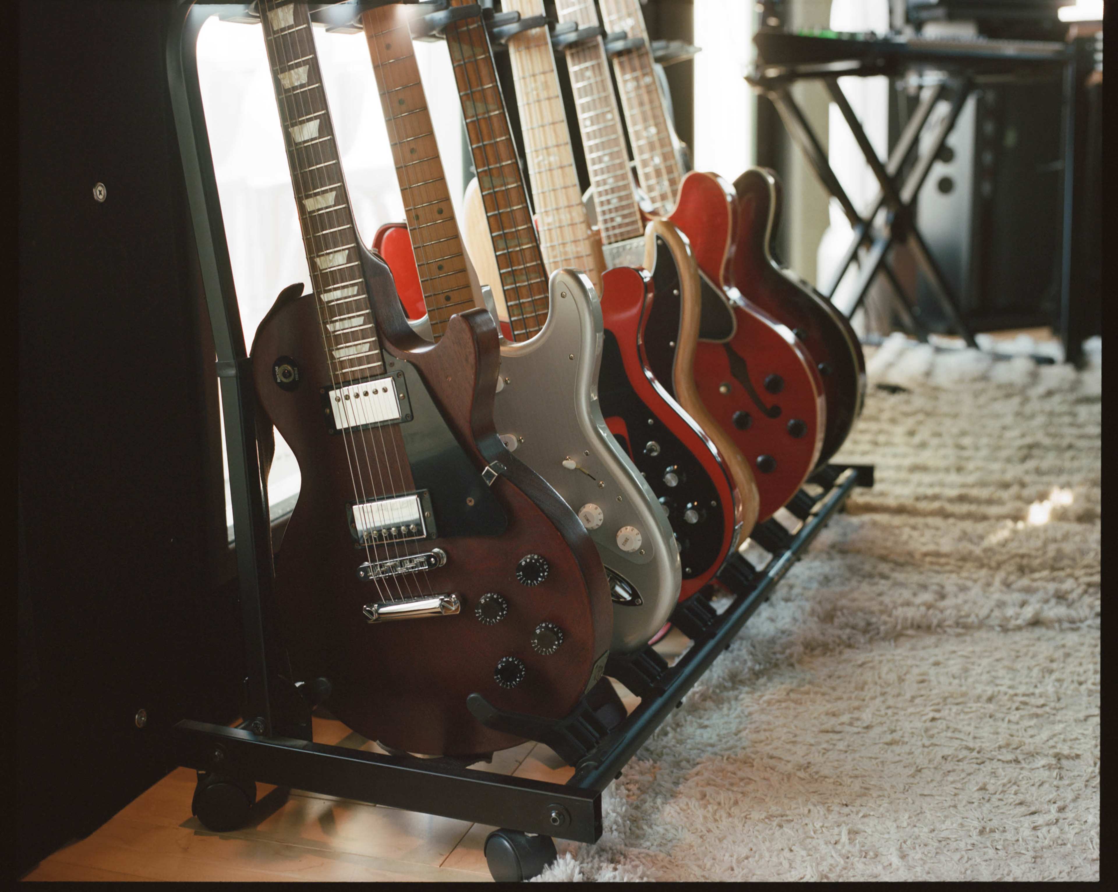 A row of six electric guitars is displayed on a rack in a well-lit room with a soft rug underneath.