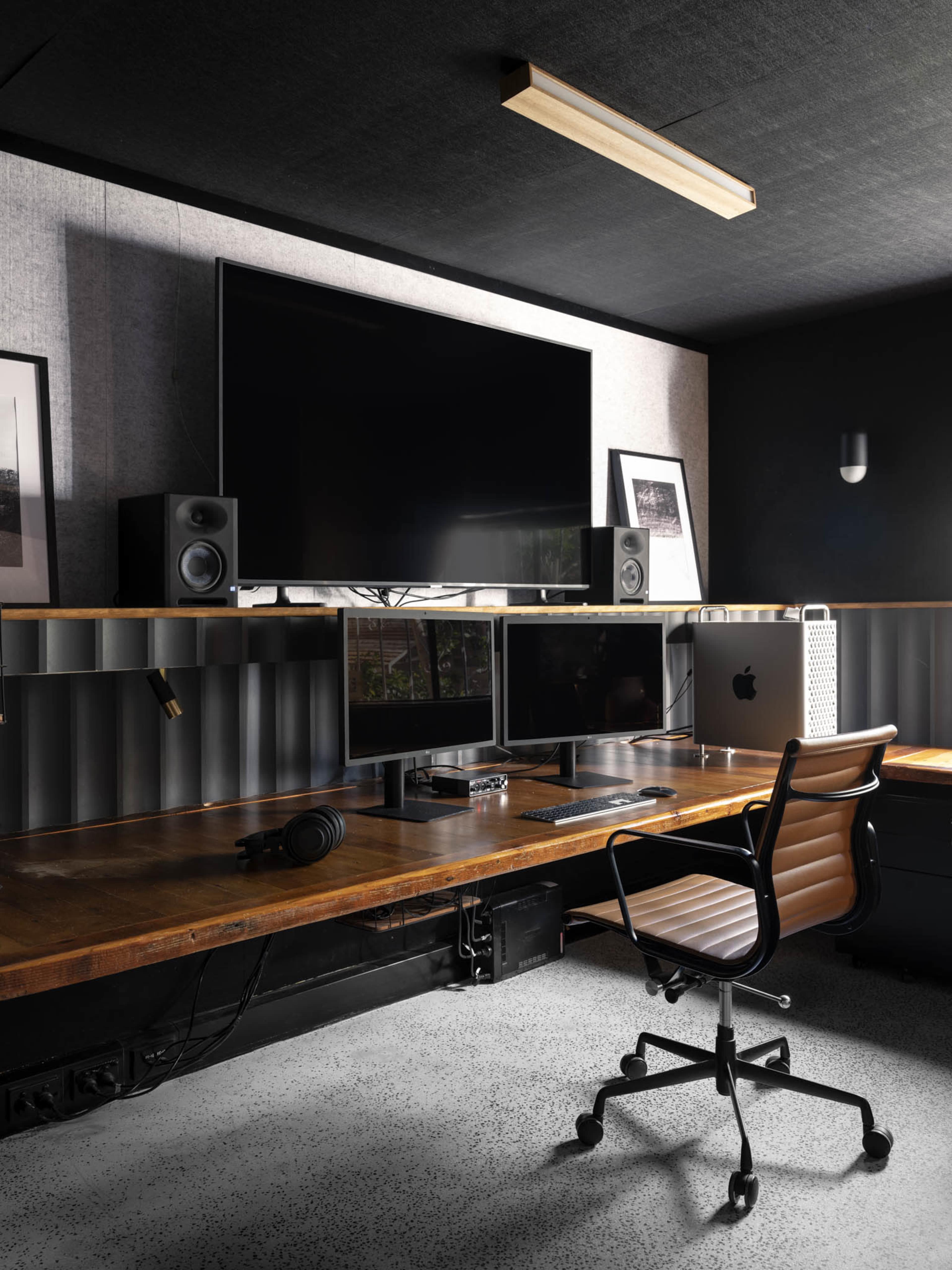 The image shows a modern workspace featuring a large television, two computer monitors, a sleek wooden desk, and a rolling office chair.