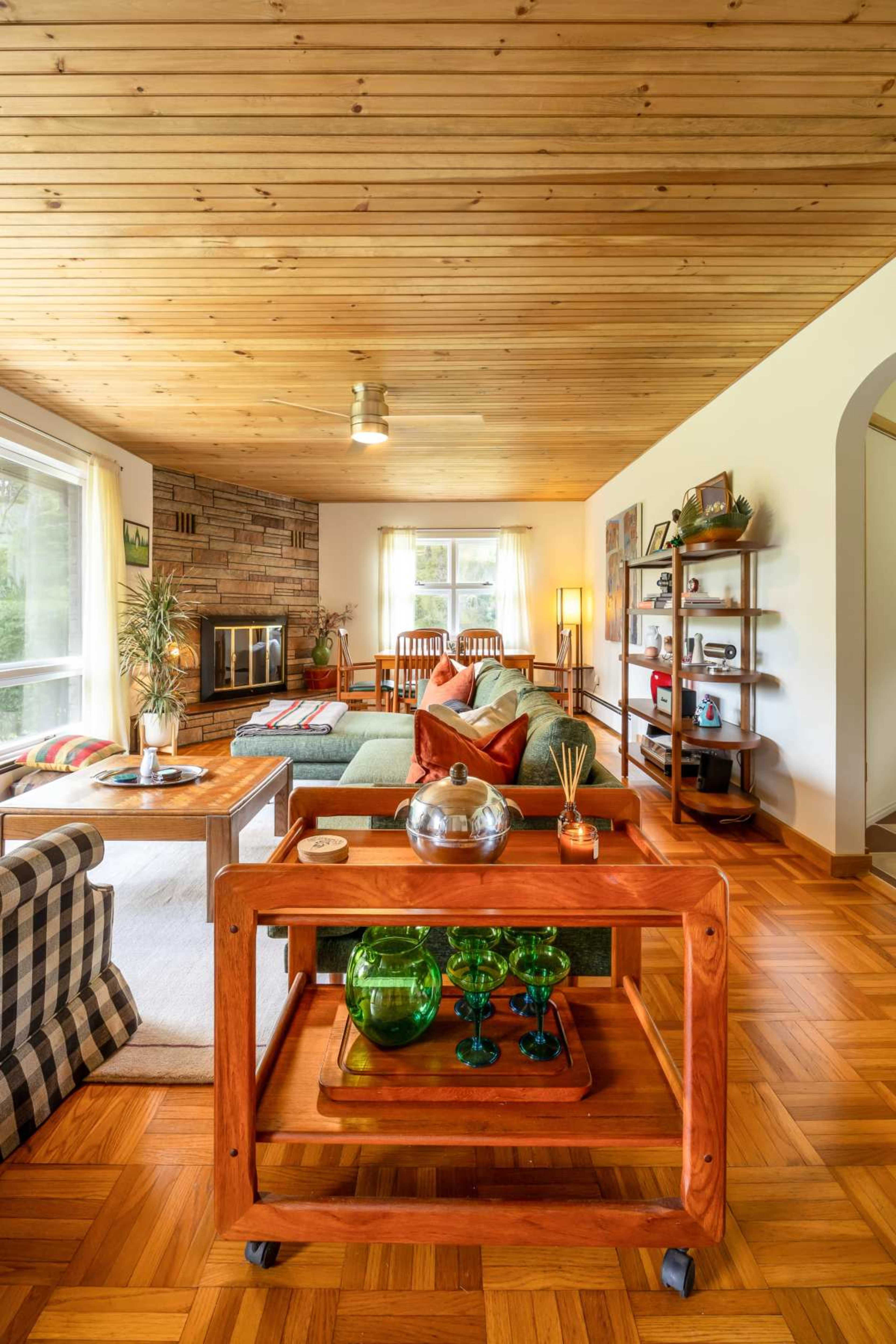 The image shows a spacious living room featuring a green sofa, a wooden coffee table, and a decorative wooden cart with glassware, alongside a large window and warm wood paneling.