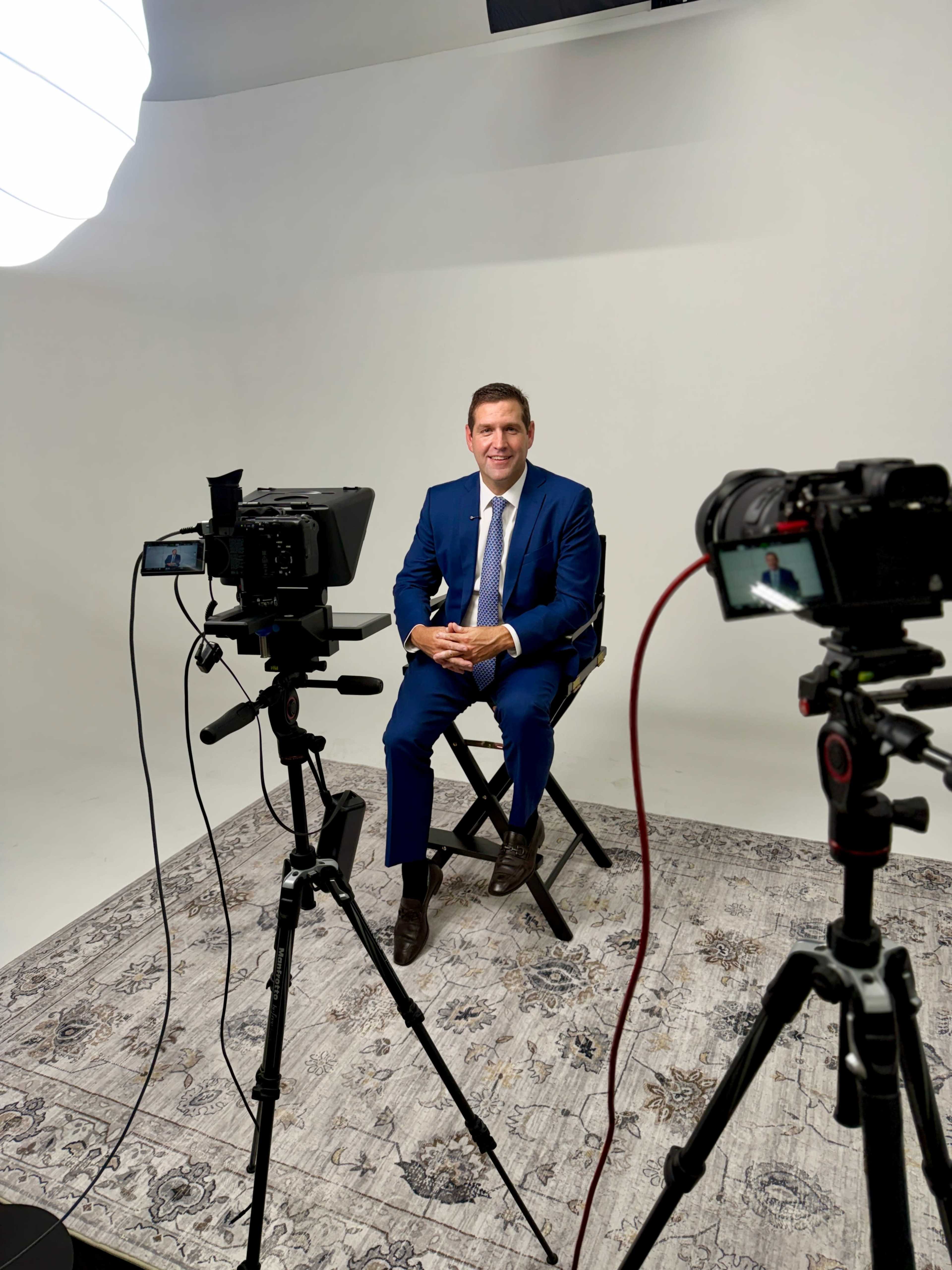 A well-dressed man sits on a stool in a studio setting, flanked by two cameras and a large softbox lighting source.