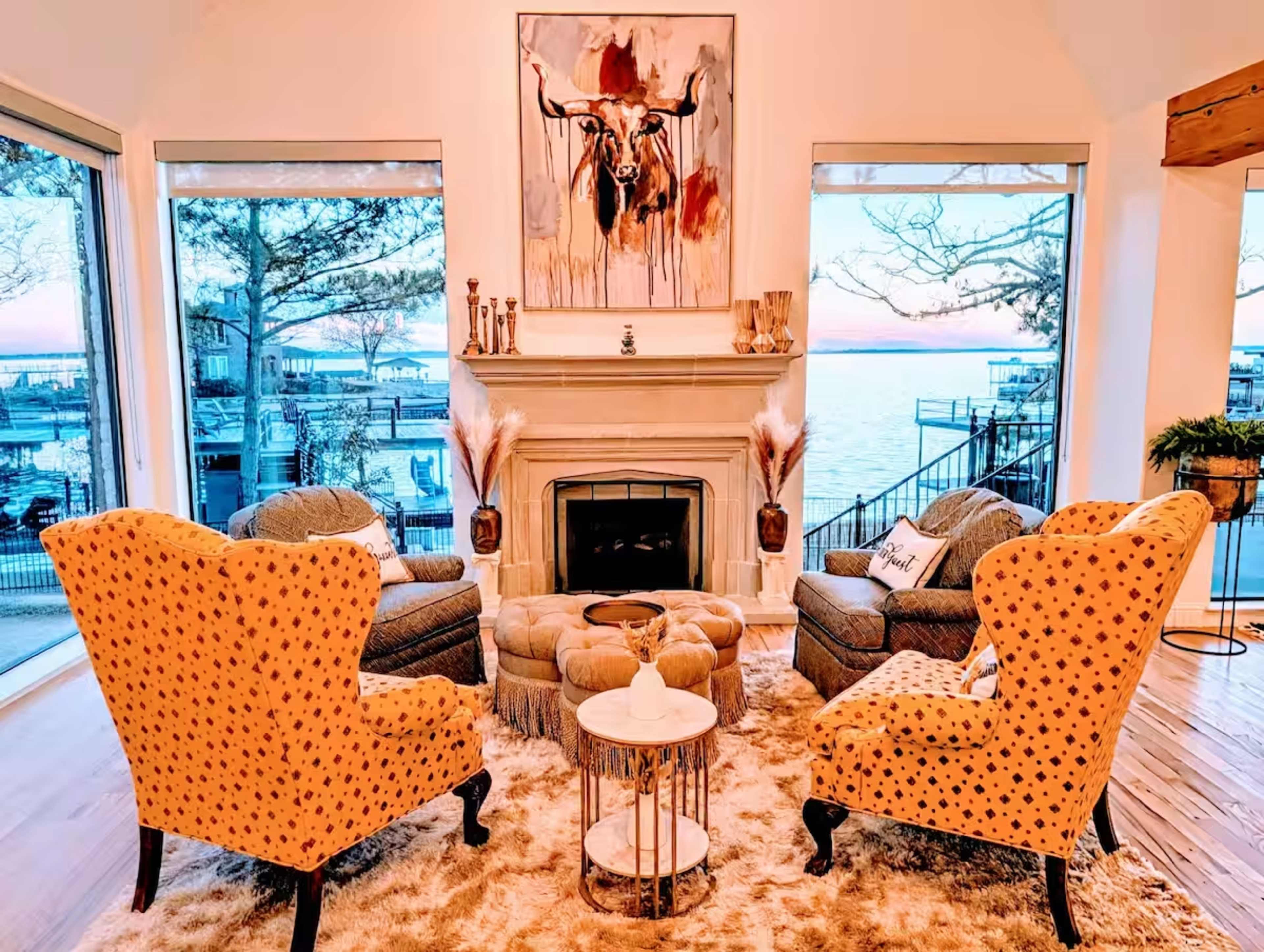 The image shows a cozy living room with two yellow upholstered armchairs, a round coffee table, and a fireplace beneath a painting of a bull, with large windows displaying a view of the water.