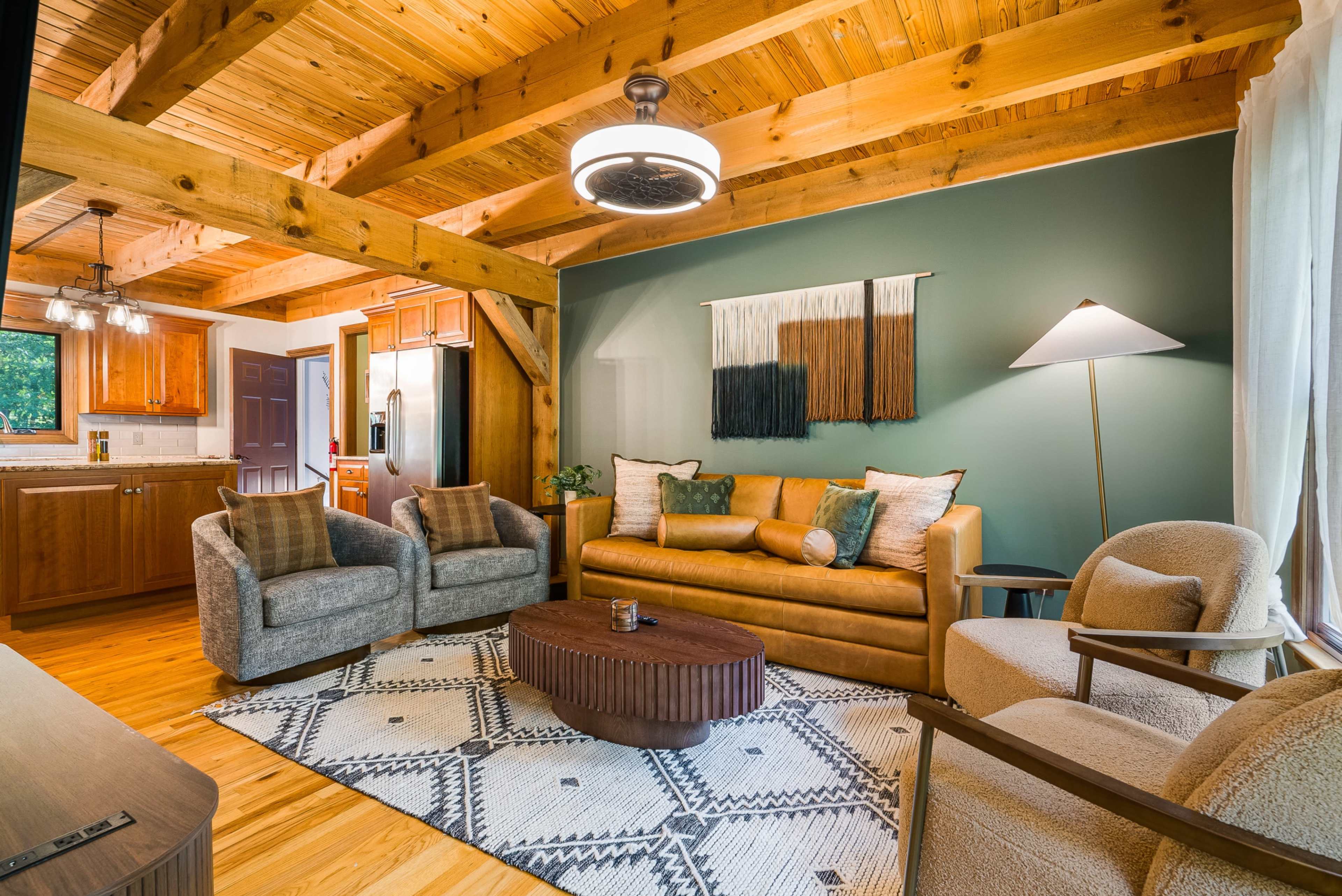 The image shows a cozy living room with wooden beams, a brown sofa, two armchairs, and a round coffee table on a patterned rug.