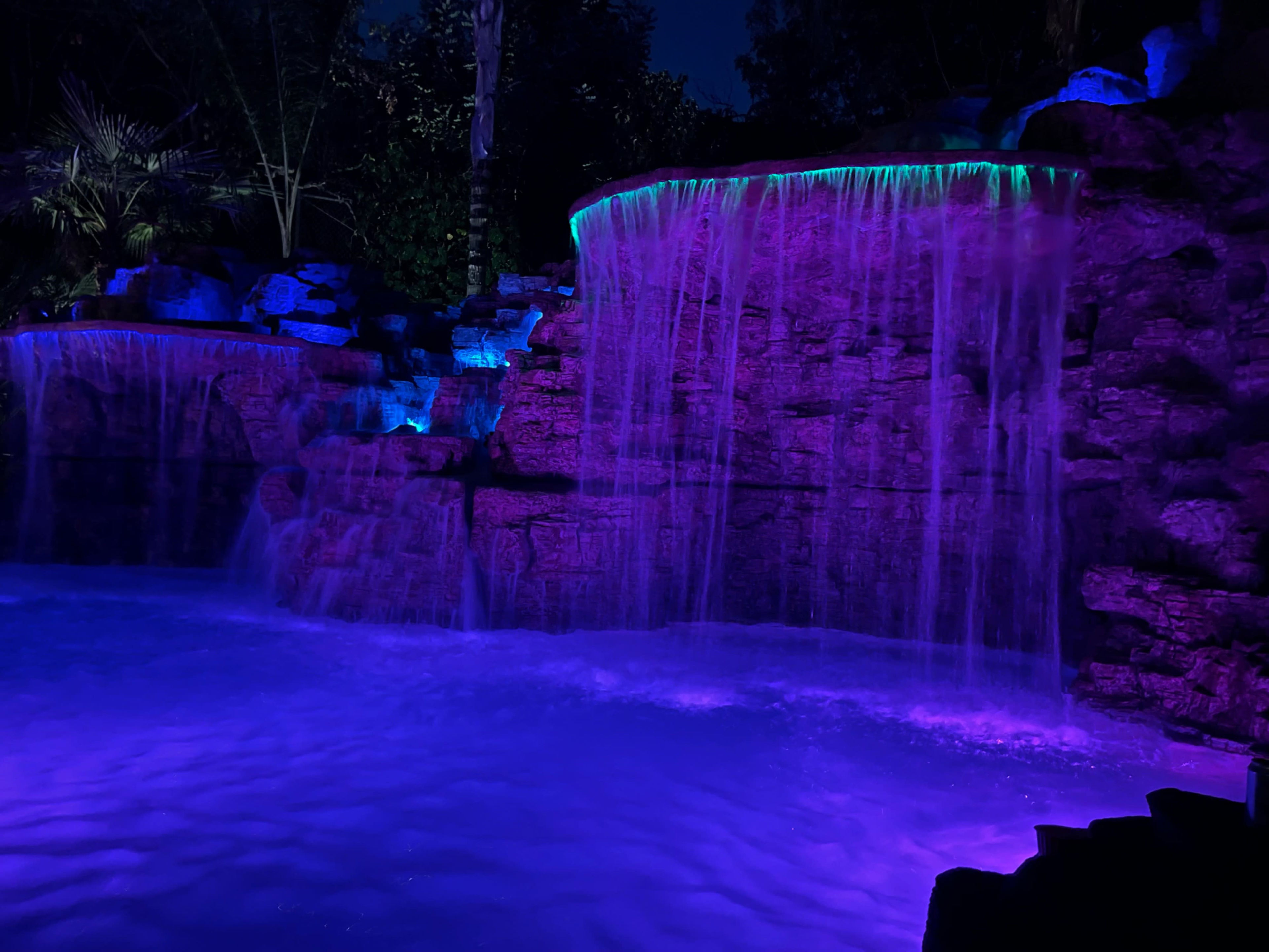 A colorful waterfall flows over rocks, illuminated by purple and blue lights at night.