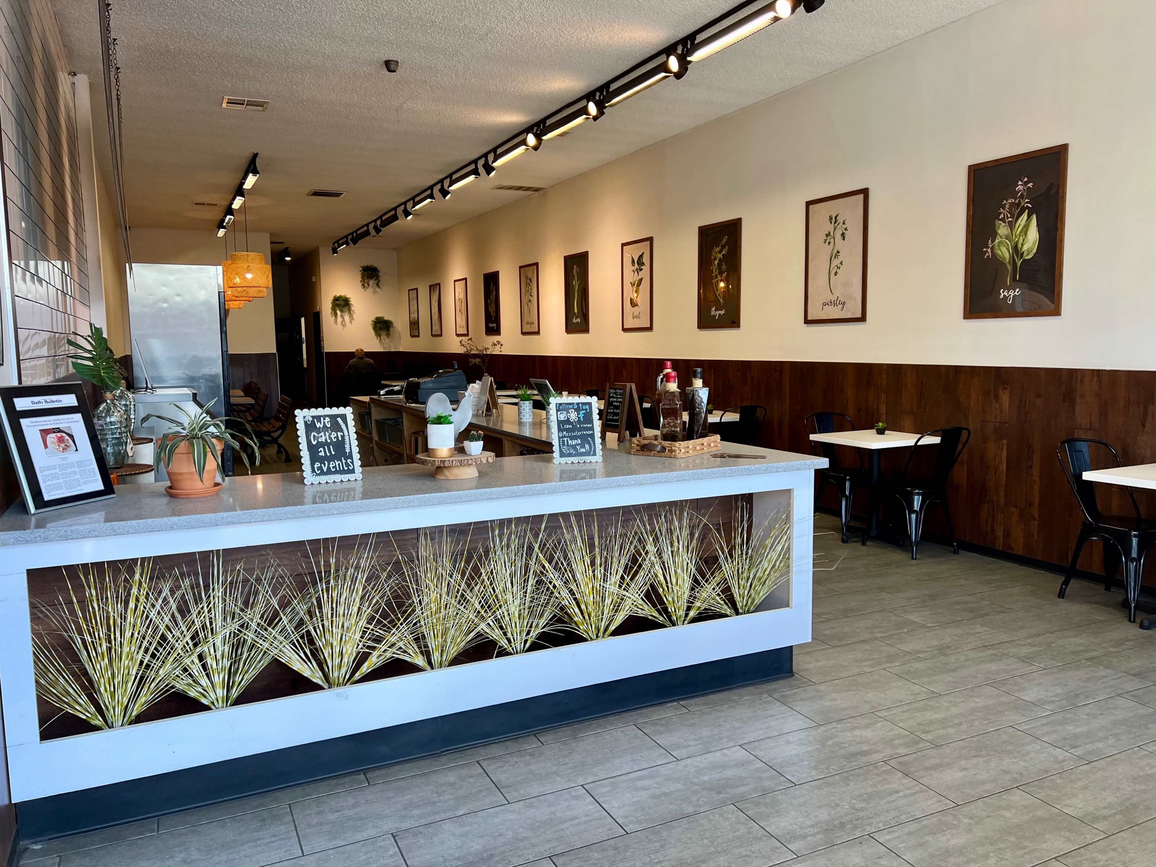 The image shows a modern café interior featuring a counter with plants, wooden accents, and framed artwork on the walls.