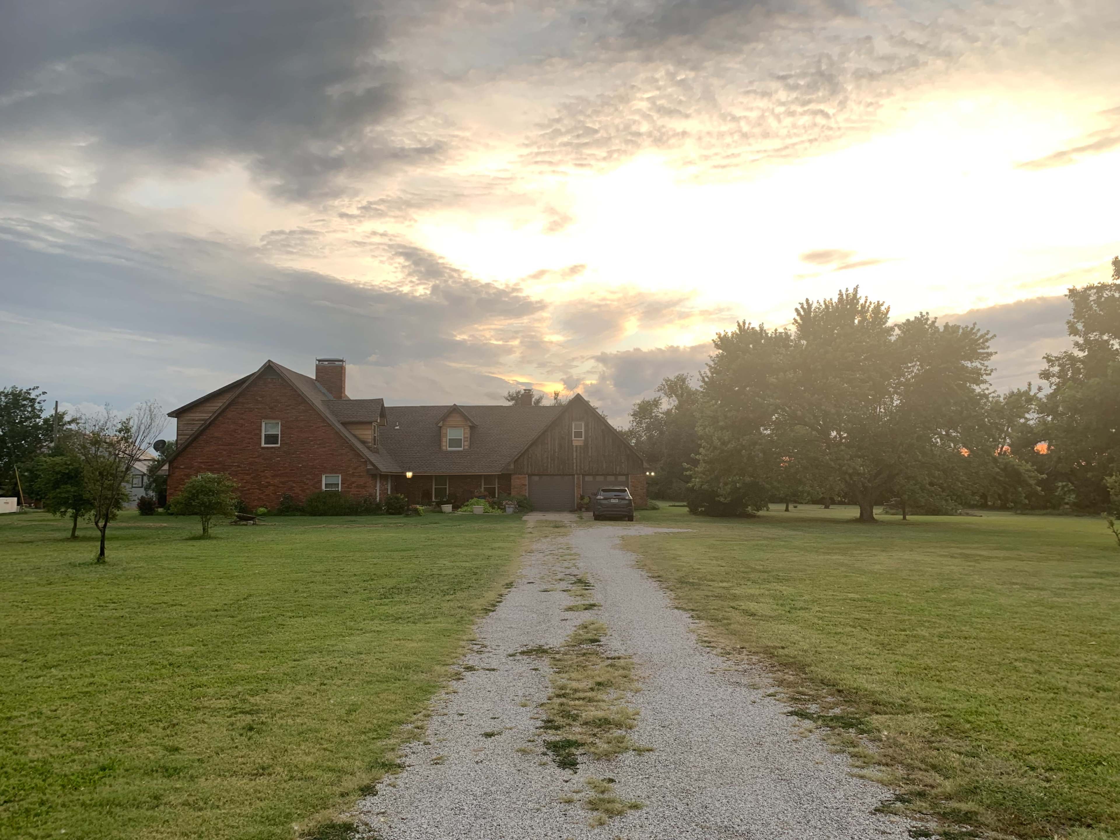 A gravel driveway leads to a brick house surrounded by trees under a cloudy sunset sky.