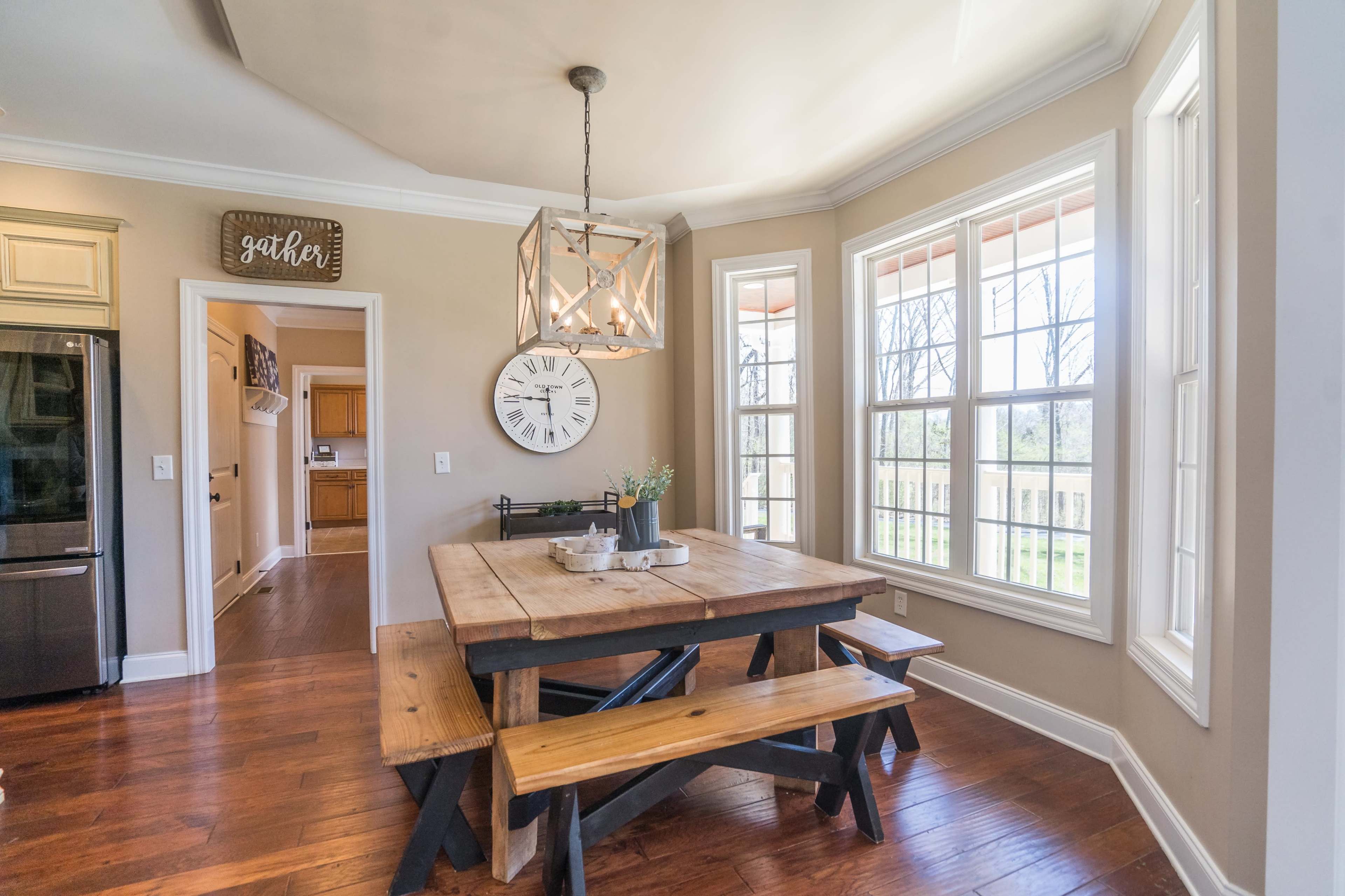 A wooden dining table with benches is situated in a brightly lit room featuring large windows and a decorative chandelier.