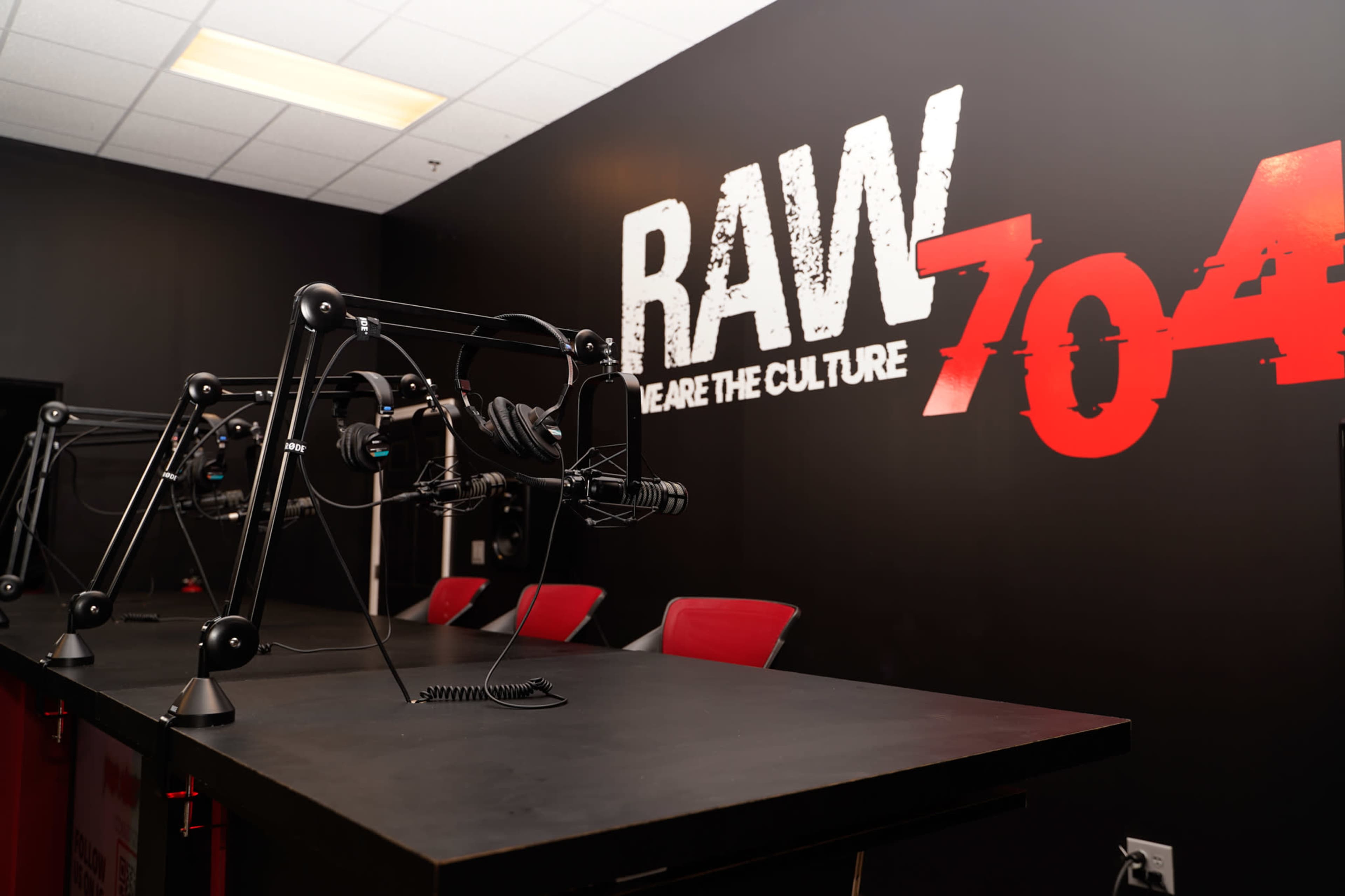 A modern radio studio with black walls, a large "RAW 704" sign, and two microphone stands positioned on a dark table.