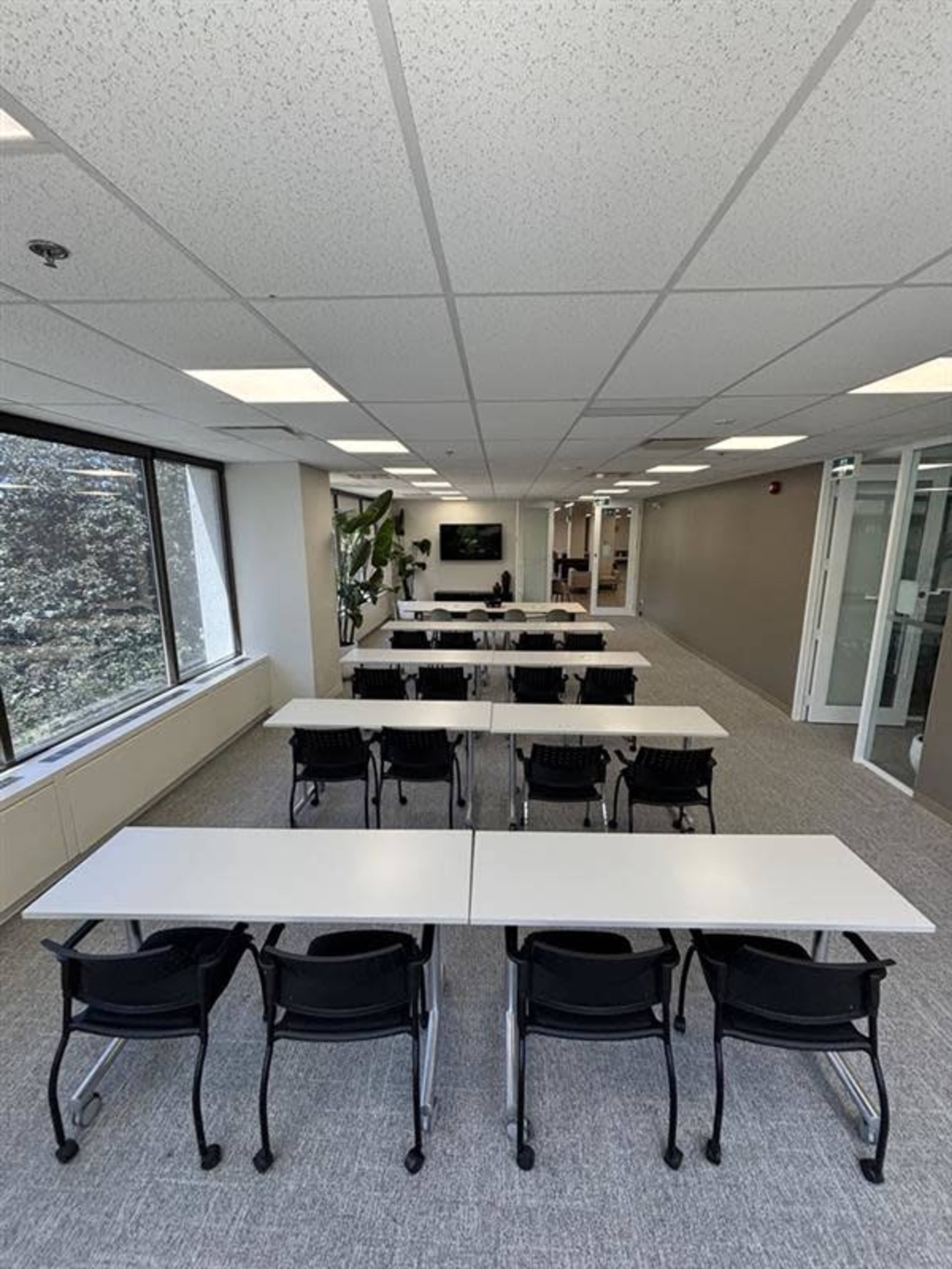 The image shows a modern, well-lit classroom or training room with several rows of tables and chairs facing a screen at the front.