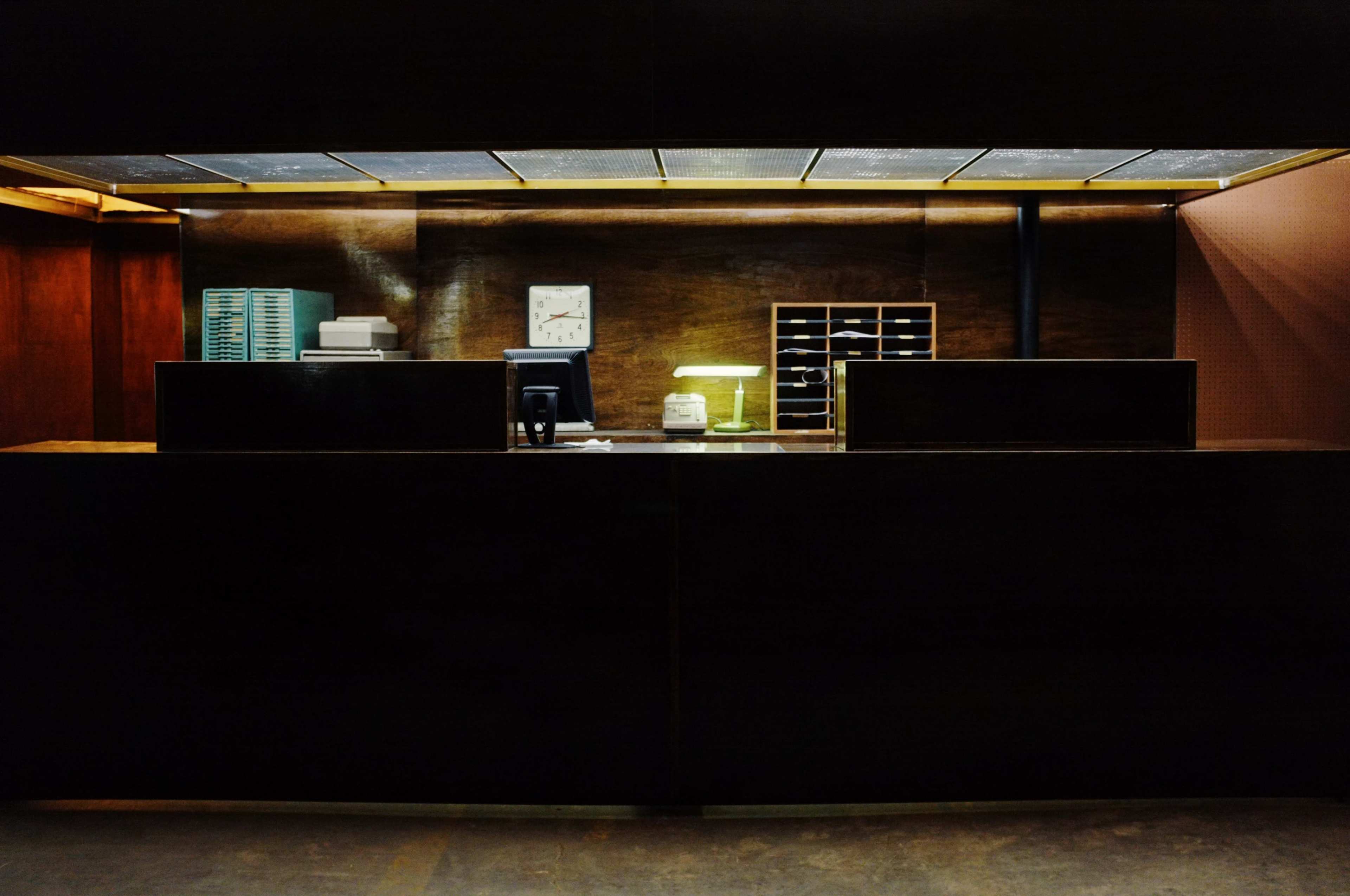 A wooden reception desk with a clock, a lamp, and a stack of items is illuminated against a dark background.