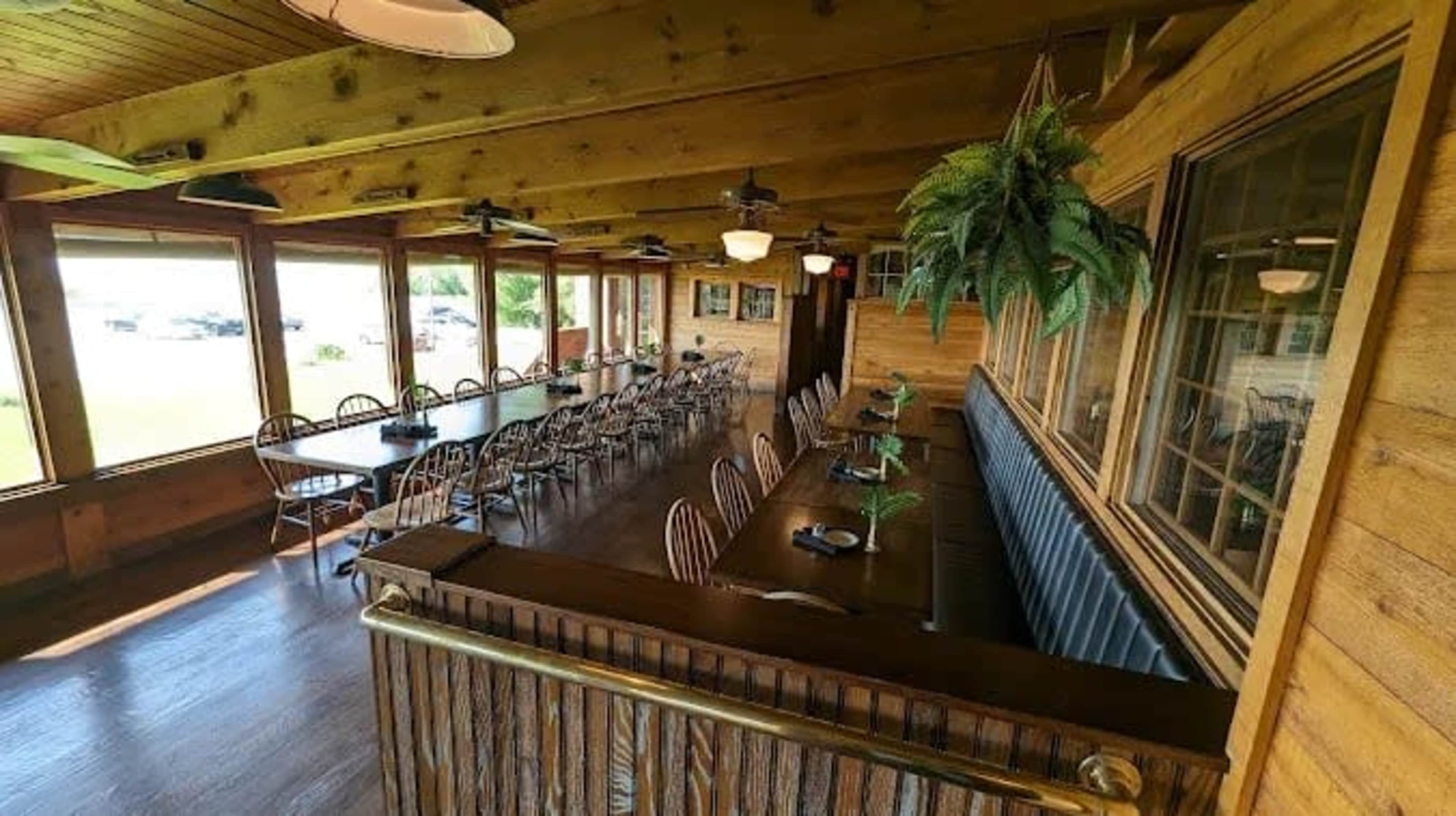 The image shows a spacious dining area with large windows, wooden walls, and multiple tables arranged in a row, suitable for gatherings.