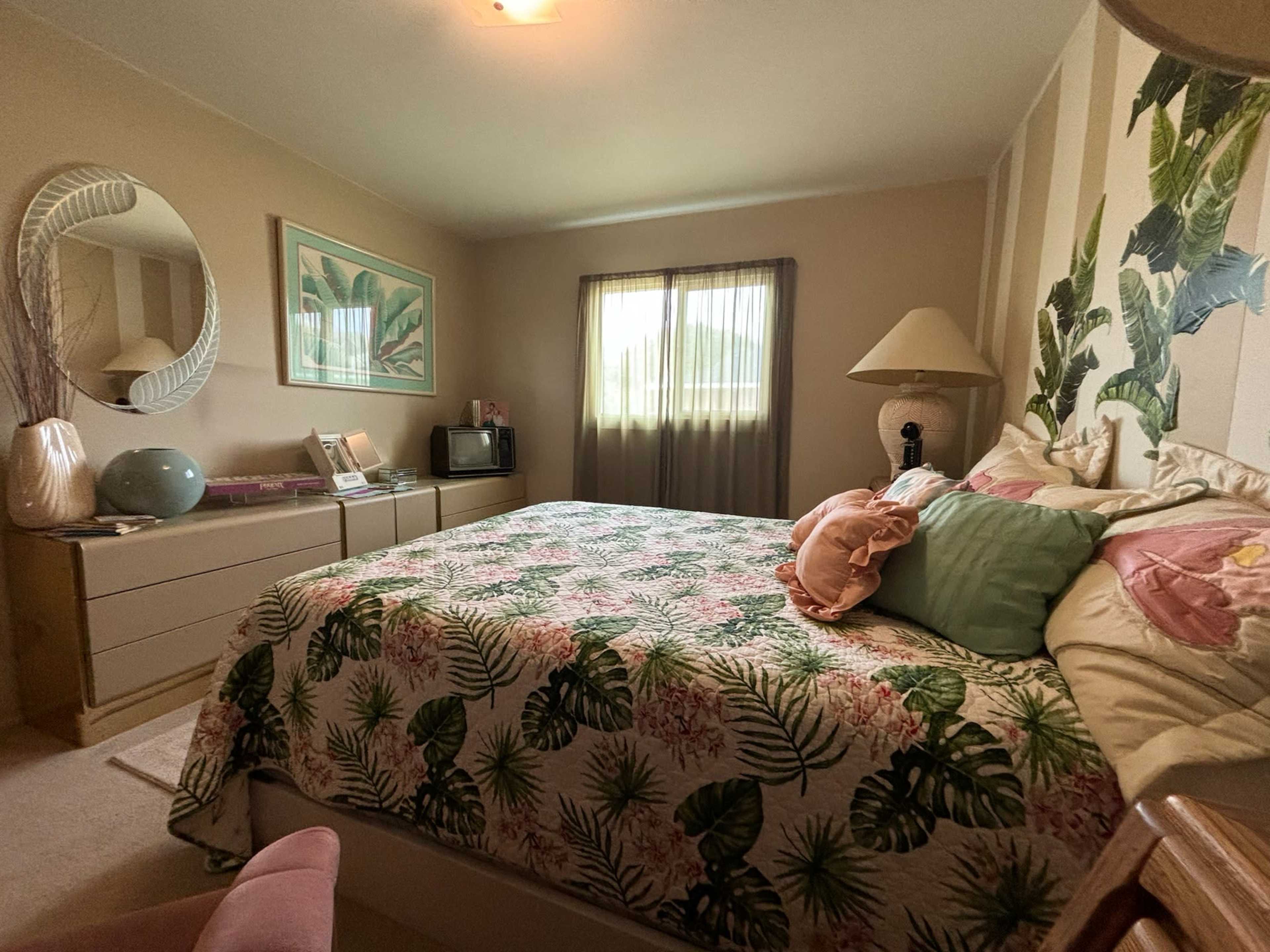 The image shows a well-decorated bedroom featuring a large bed with a floral-patterned duvet, a lamp, a mirror, and a dresser, all arranged under natural light from a window with sheer curtains.