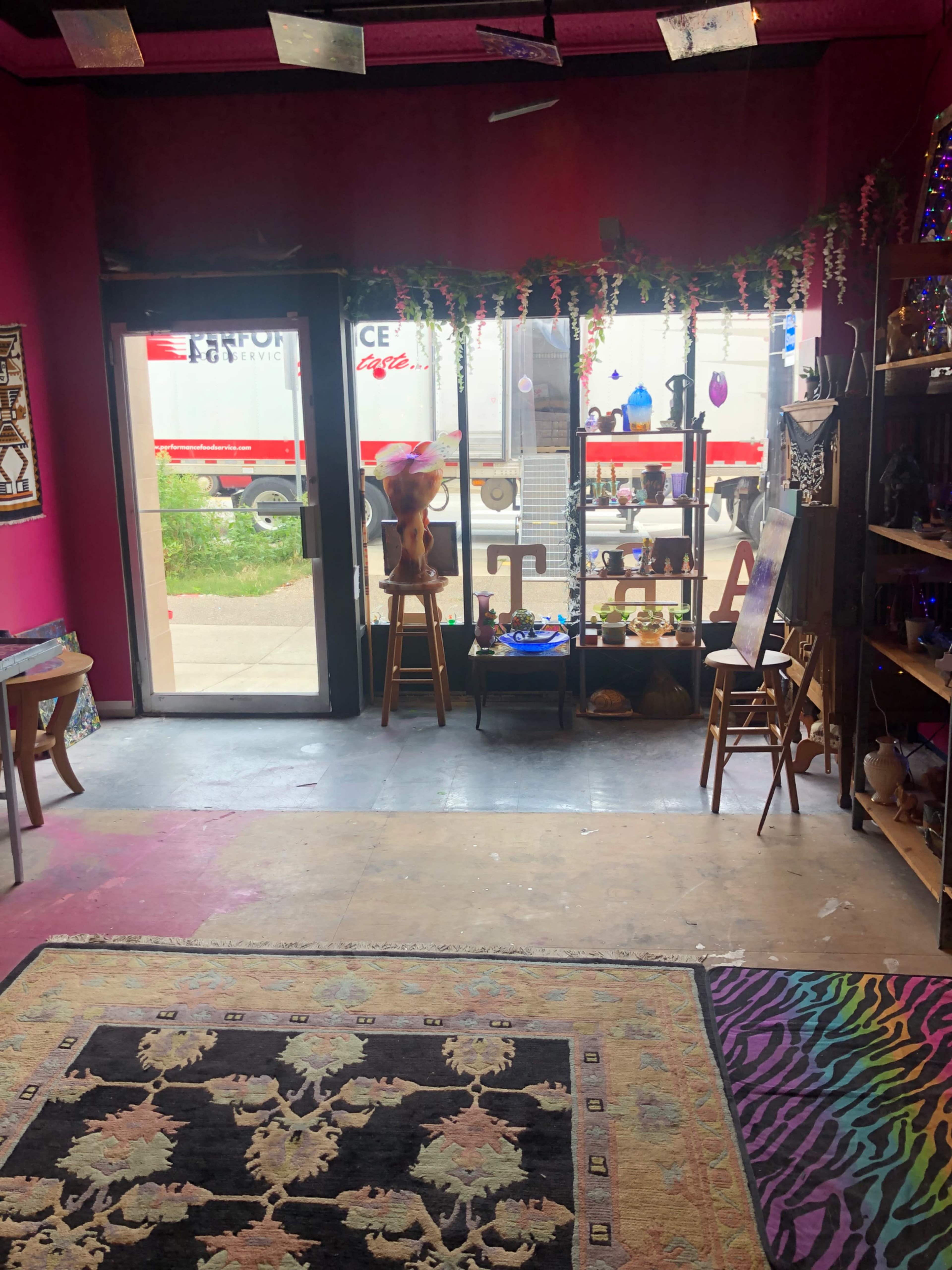 A vibrant interior of a shop with colorful decor, a patterned rug, shelves displaying various items, and a glass door leading to a view of a truck parked outside.