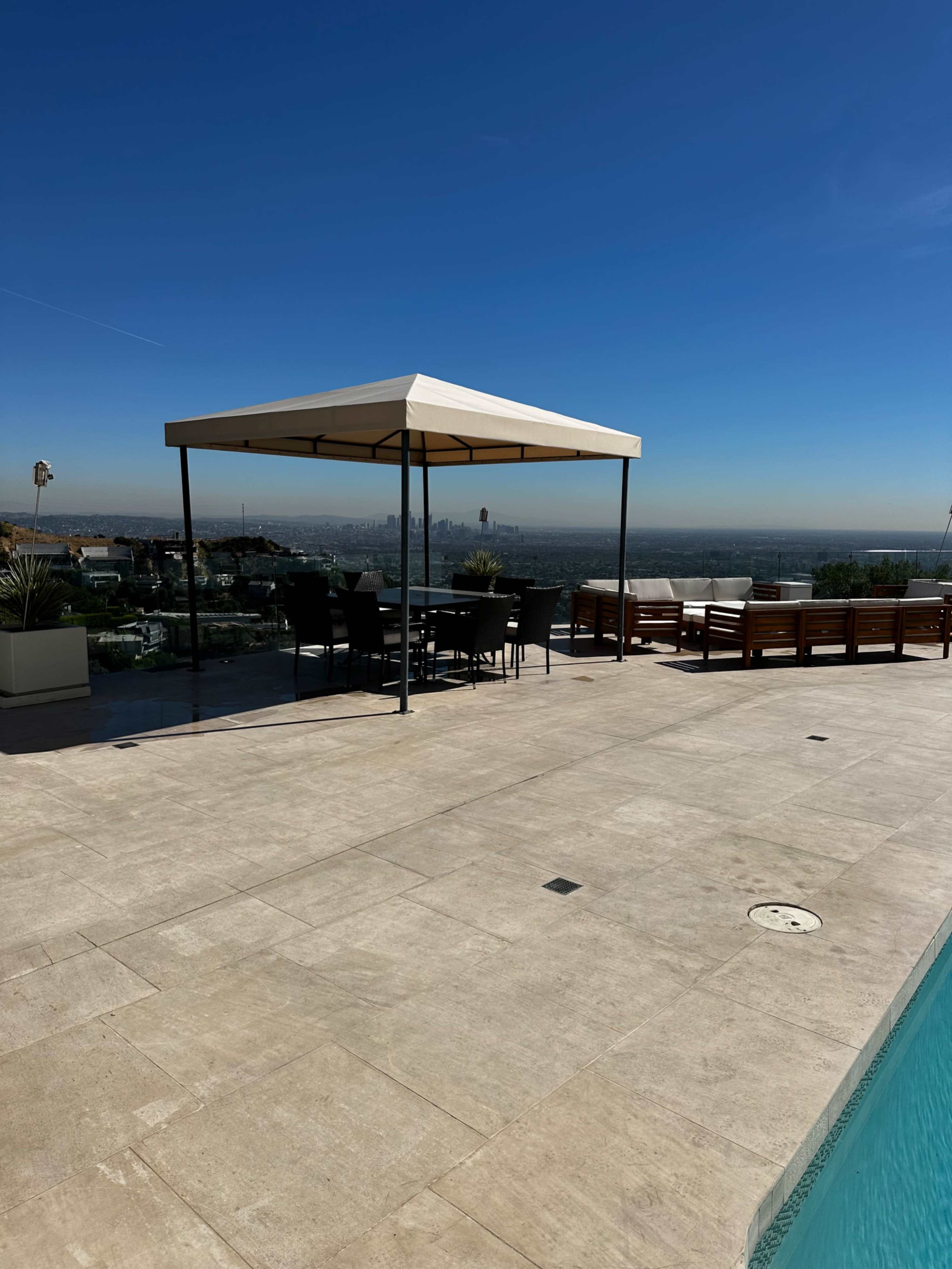 A shaded patio area with black chairs and a table overlooks a pool and city skyline.