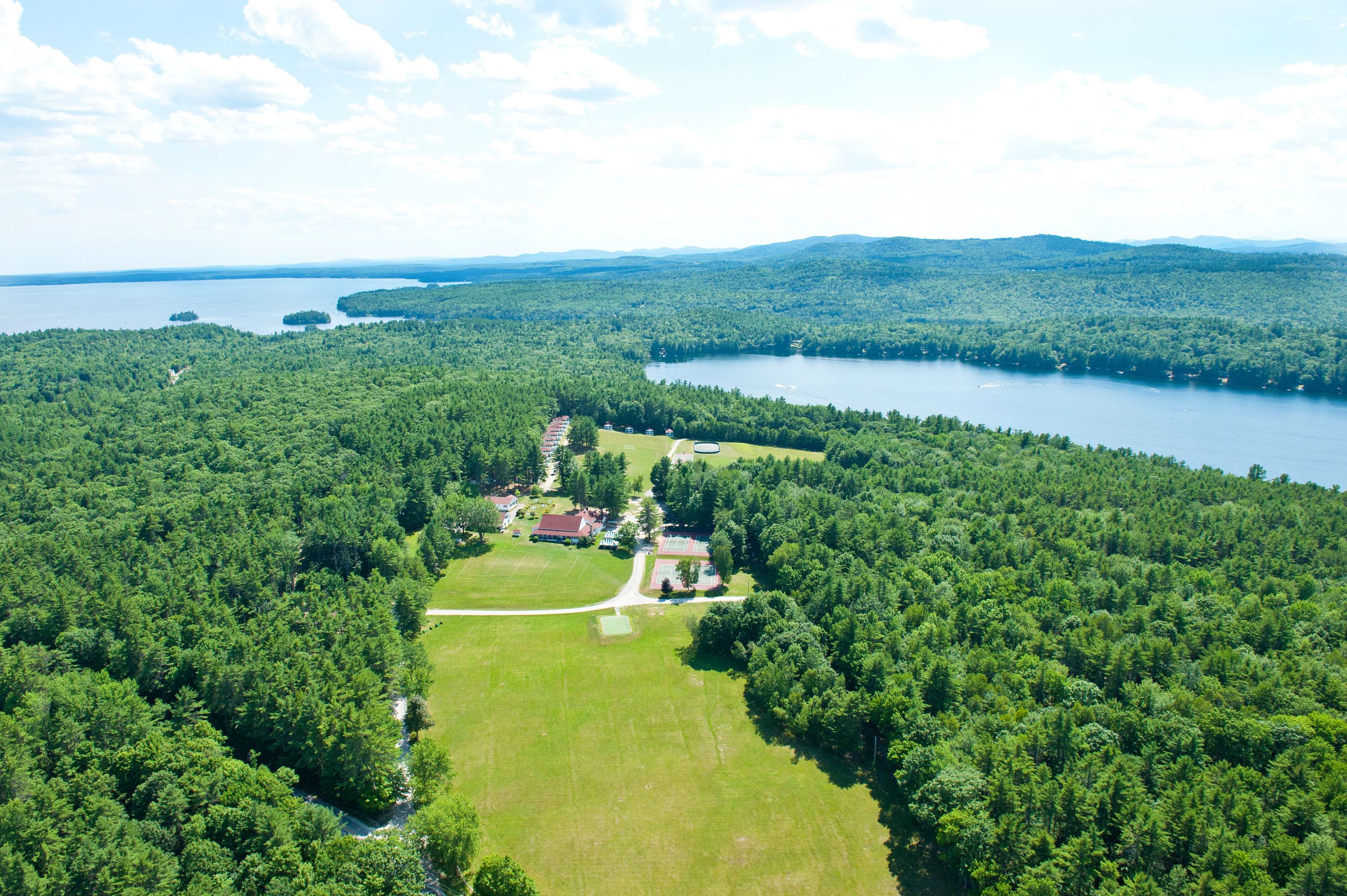 An aerial view shows a lush green landscape with a large lake on the left and a river on the right, dotted with a few buildings amidst dense trees.