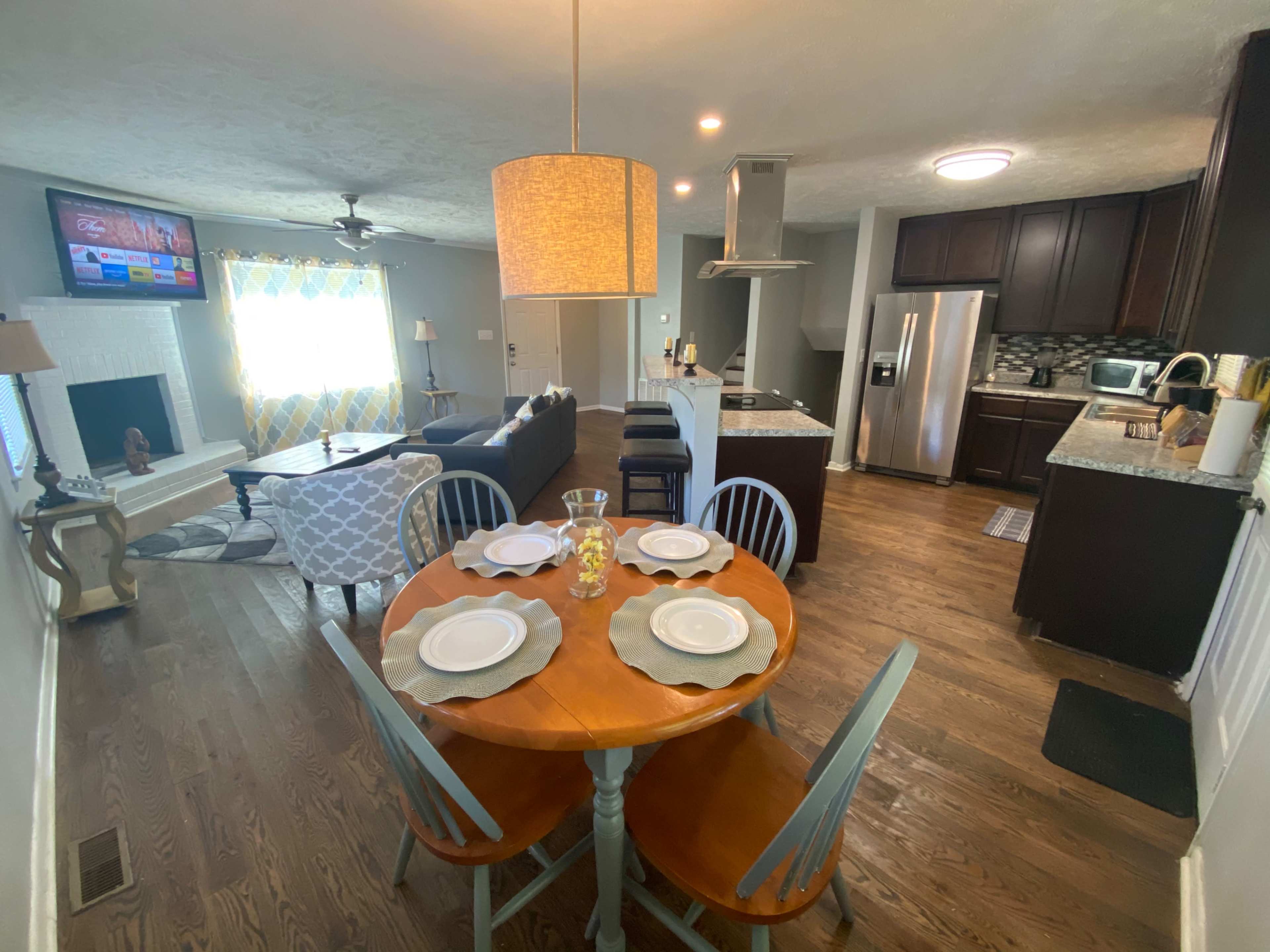A modern open-concept living space featuring a dining area with a round table and chairs, a furnished living room with a television, and a kitchen with granite countertops.
