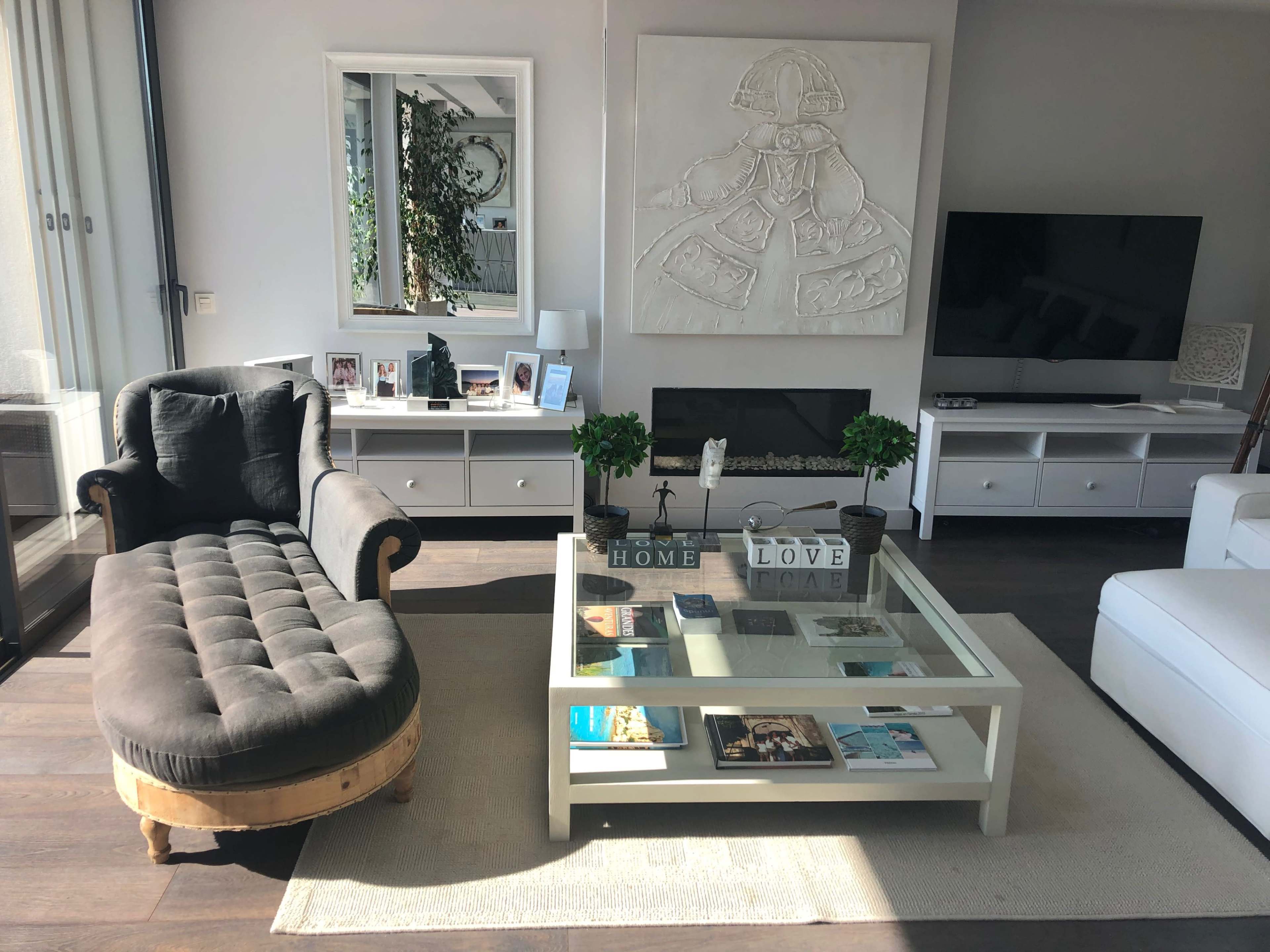 The living room features a chaise lounge, a glass coffee table stacked with books, a flat-screen TV, and a large wall art piece.