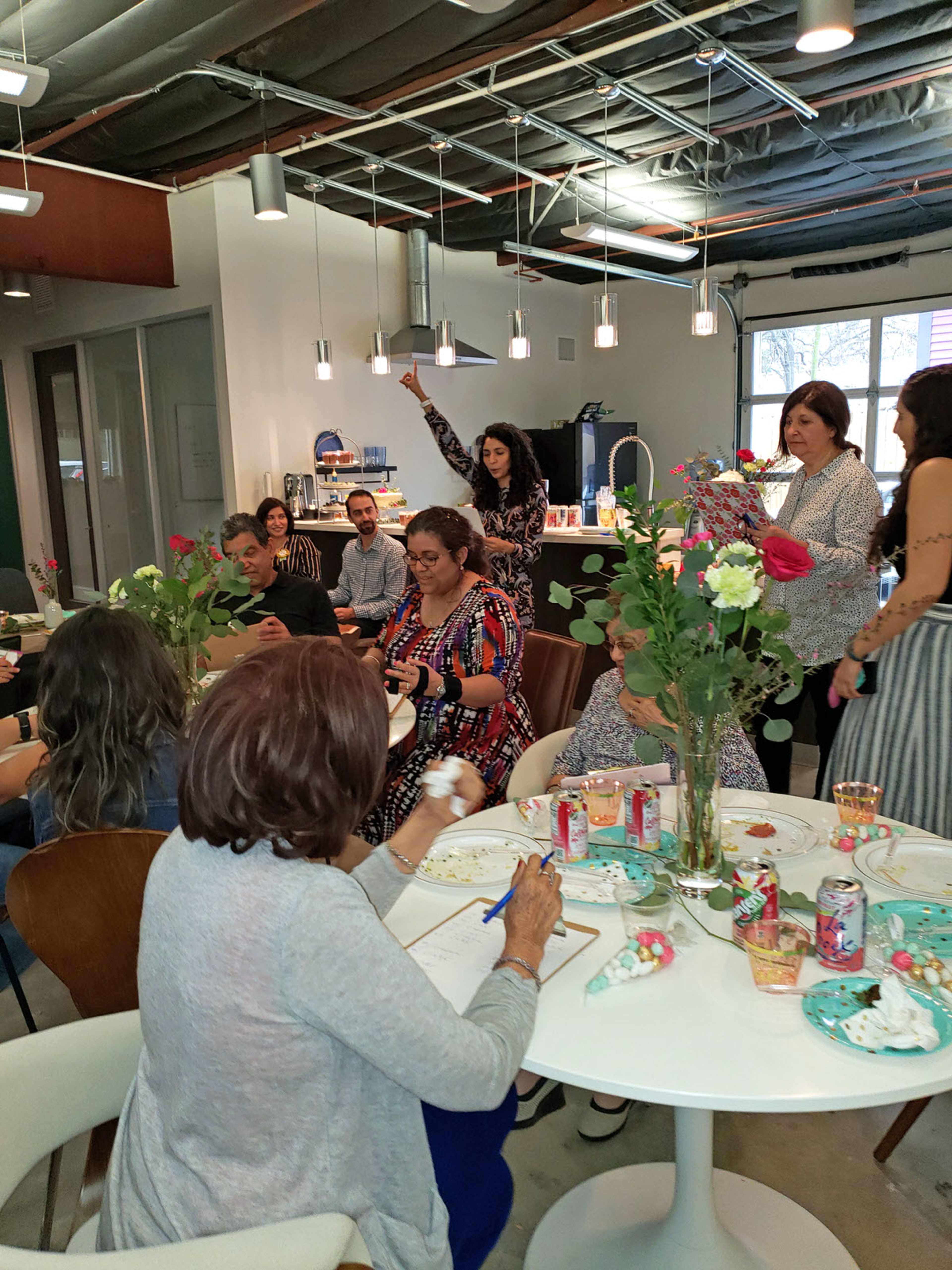 A group of people are gathered around a table in a brightly lit room, engaged in crafting activities with flowers and refreshments.