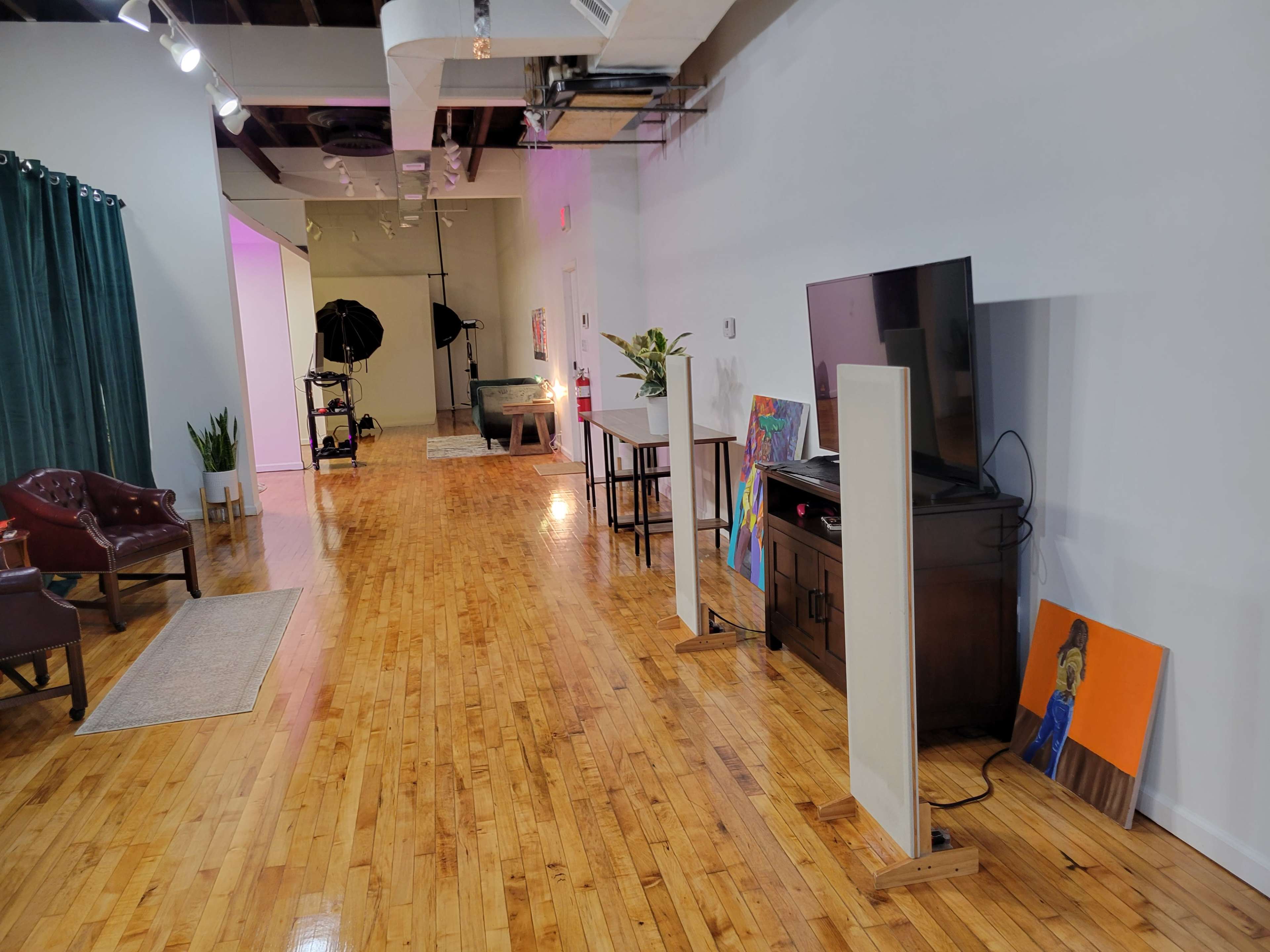 The image shows a spacious room with polished wooden floors, featuring a seating area with a chair, a television on a furniture unit, and various artworks leaning against the wall.