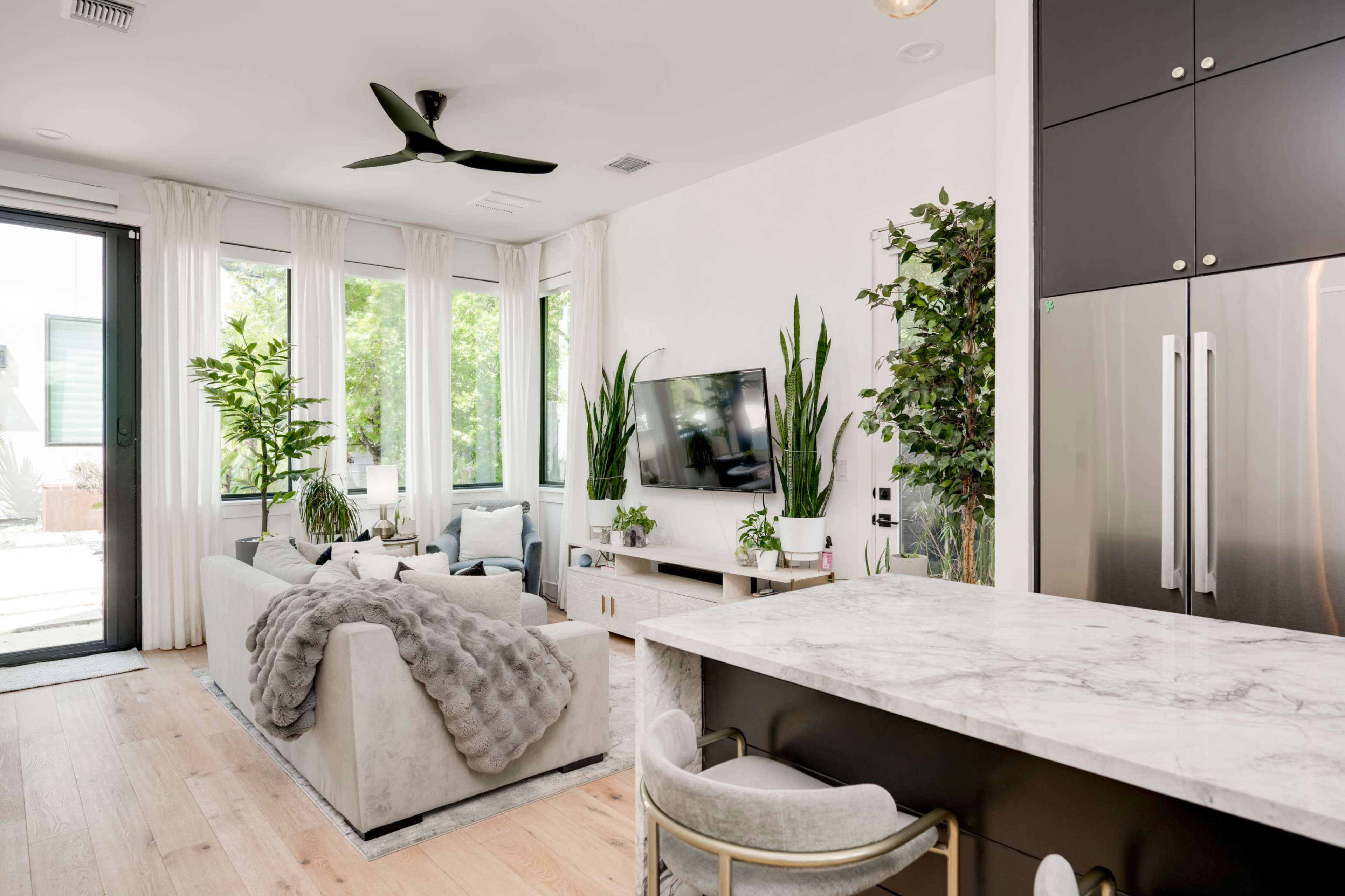 The image shows a modern living room with a large window, plants, a television mounted on the wall, and an adjacent kitchen island with seating.