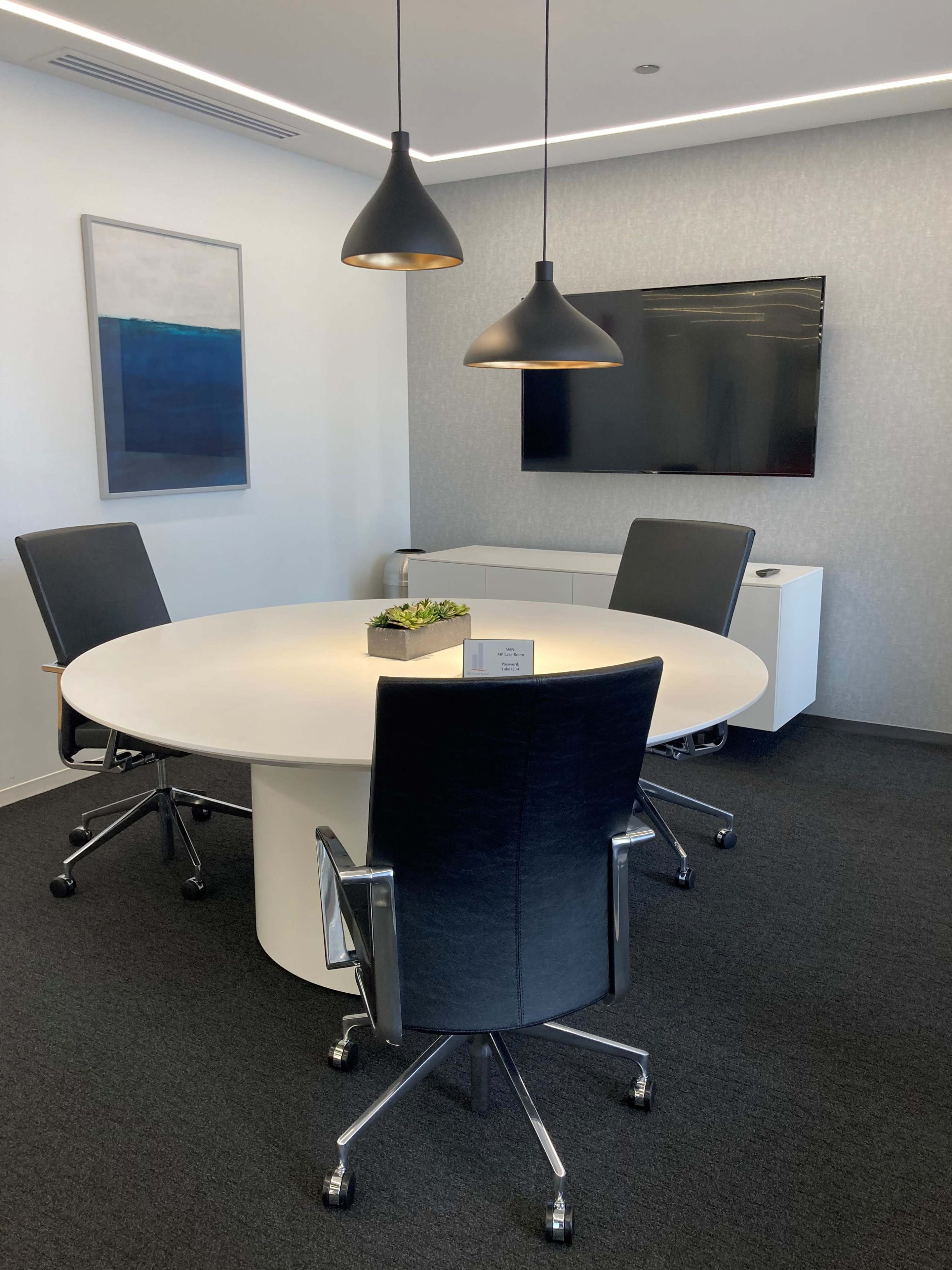 The image shows a modern meeting room featuring a round table surrounded by three black leather chairs, with a wall-mounted television and pendant lights above.