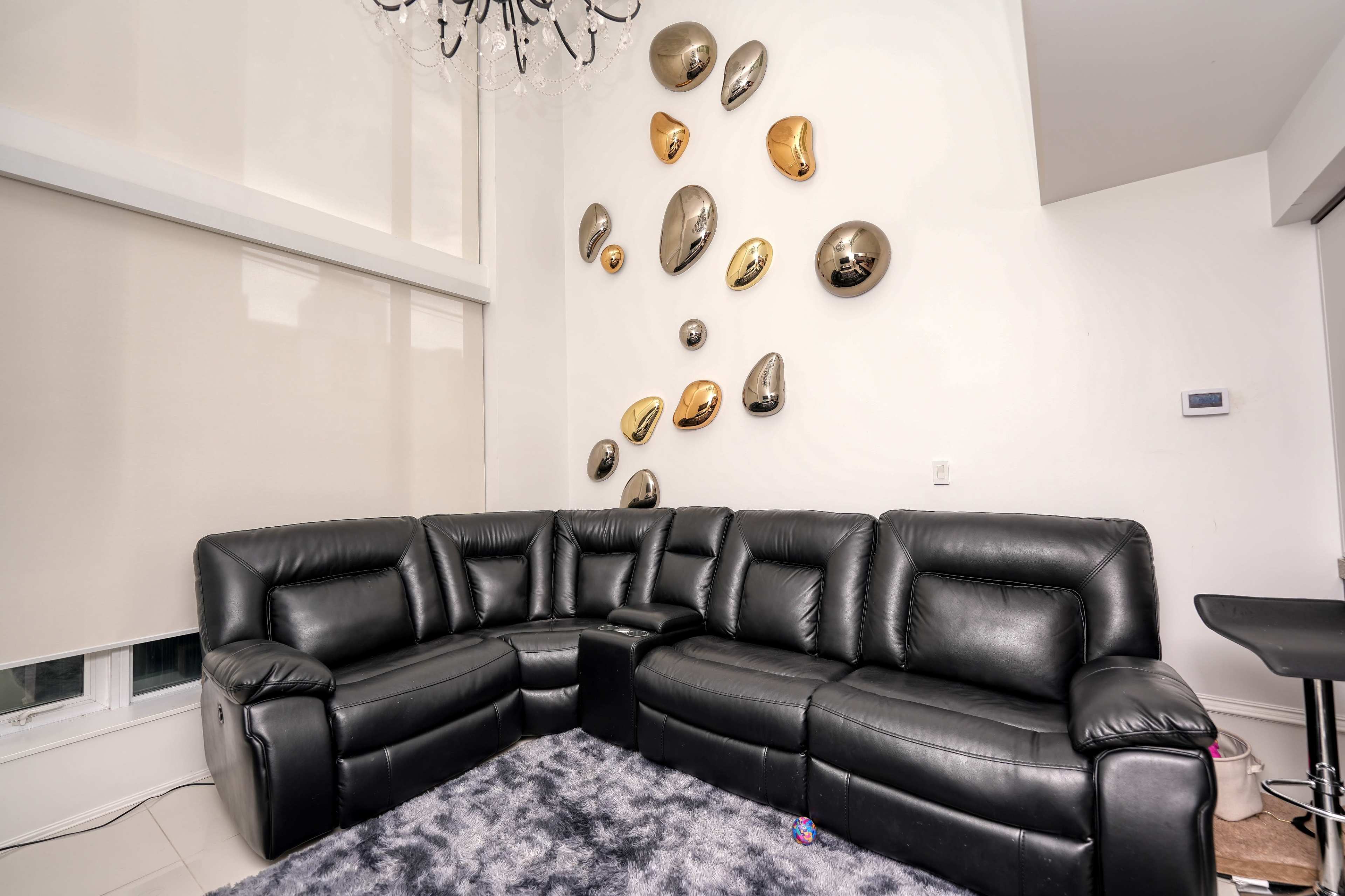 The image shows a modern living room with a black sectional sofa, a light-colored wall adorned with decorative metallic wall art, and large windows covered with beige blinds.