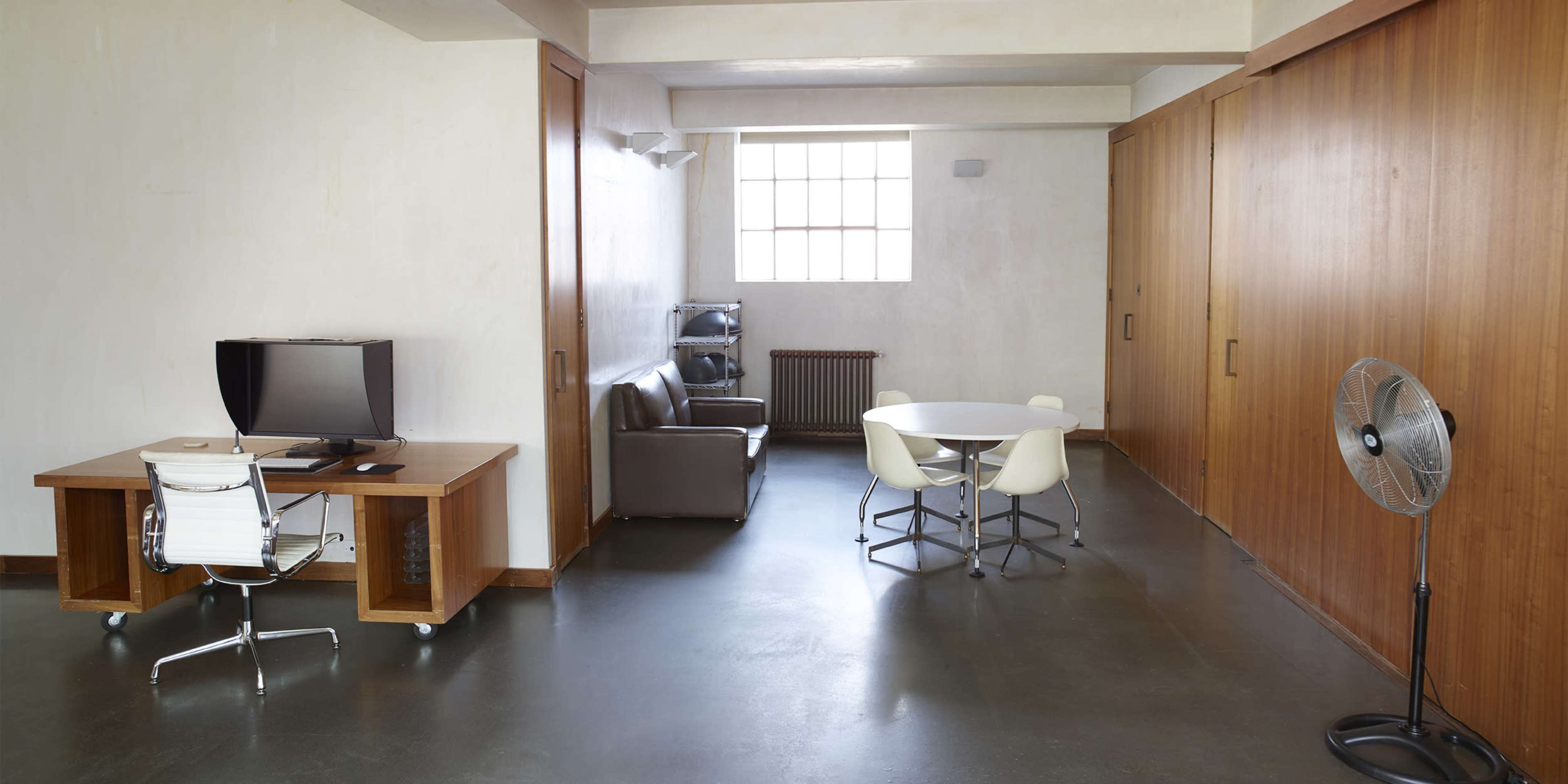 The image shows a minimalist room with a desk, television, brown sofa, round white dining table, and a standing fan.