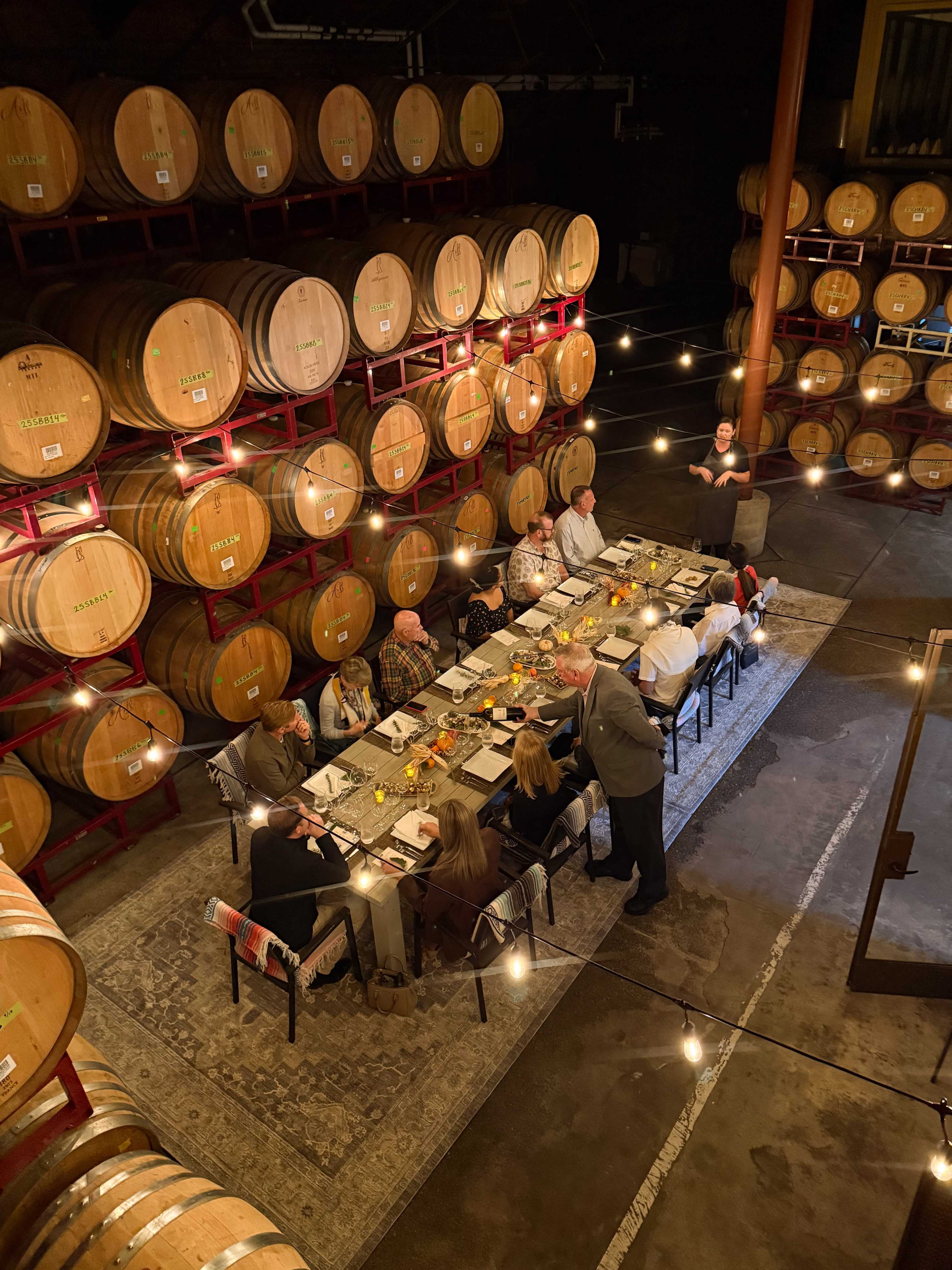 A long dining table is set for a meal in a winery, surrounded by wooden barrels and decorated with string lights.