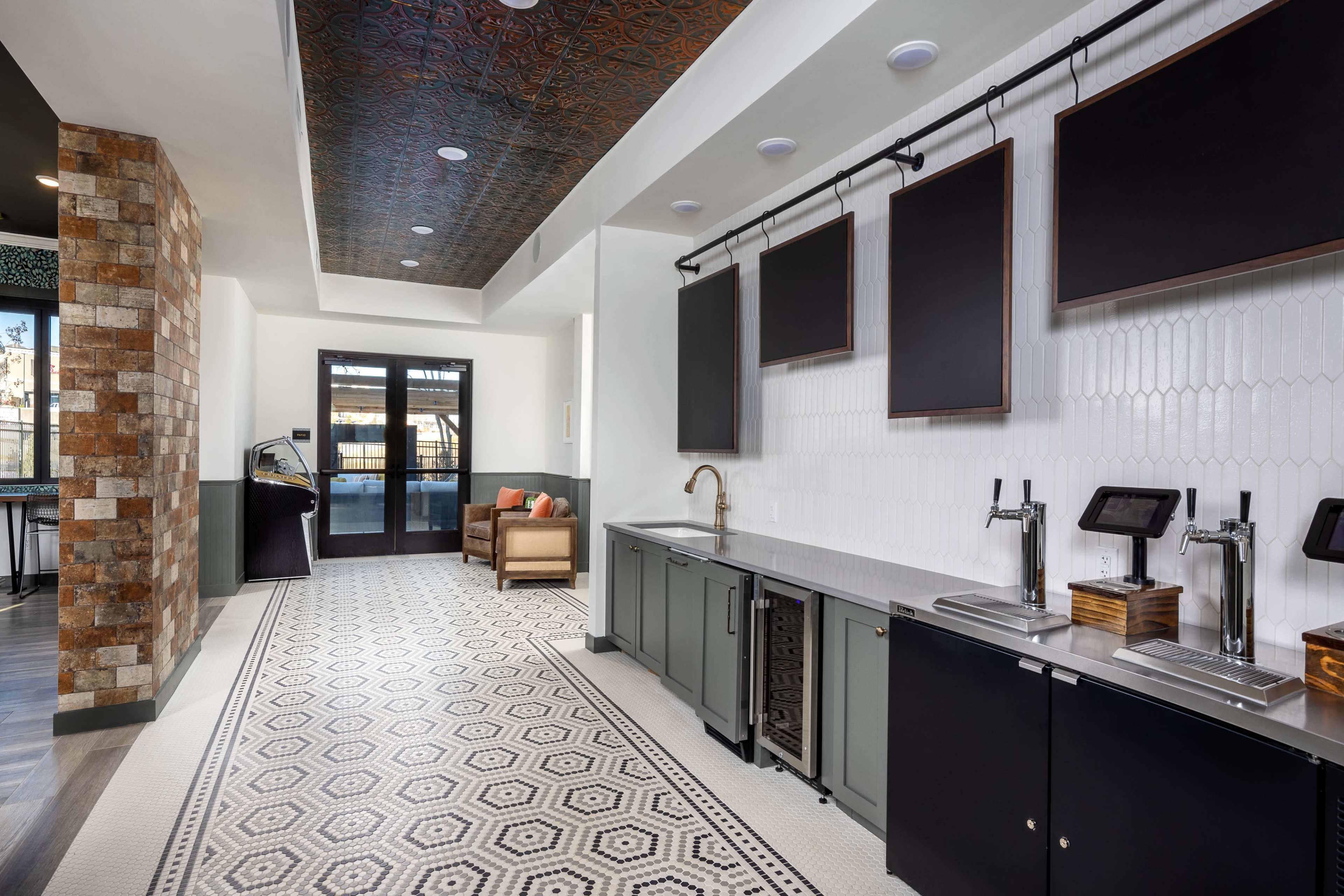 A modern kitchen area with a long countertop, taps for beverages, and a patterned floor, adjacent to a seating area.