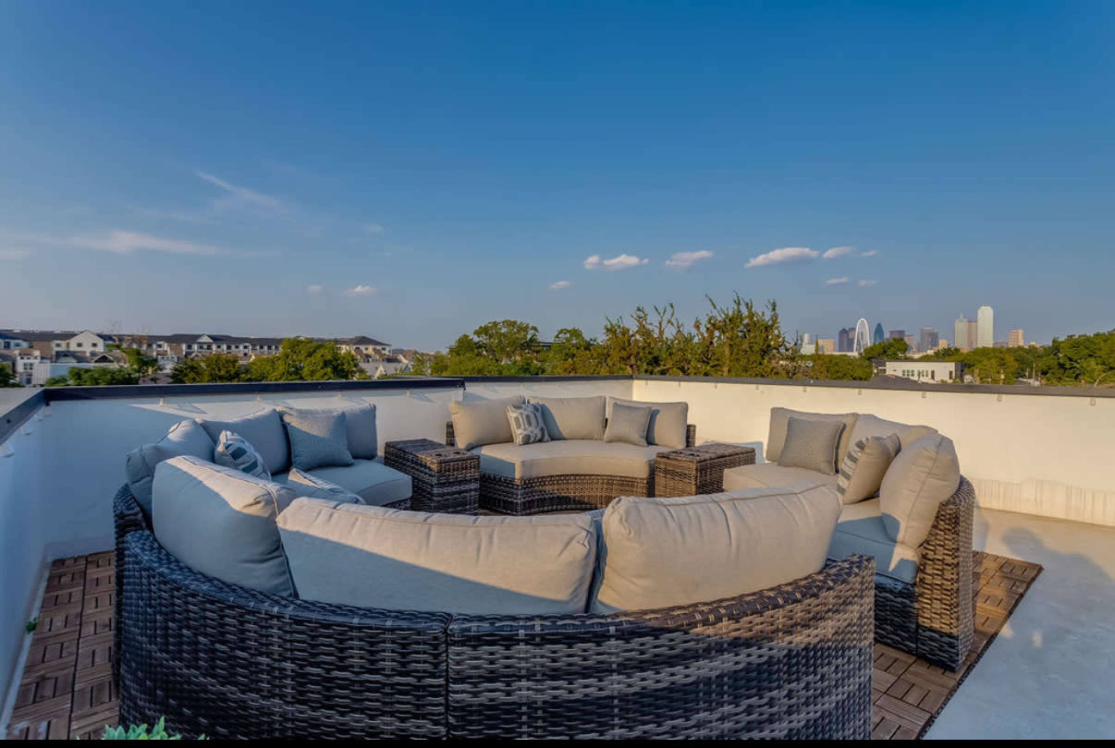 A rooftop patio features a circular arrangement of cushioned wicker seating with a view of a city skyline in the background.