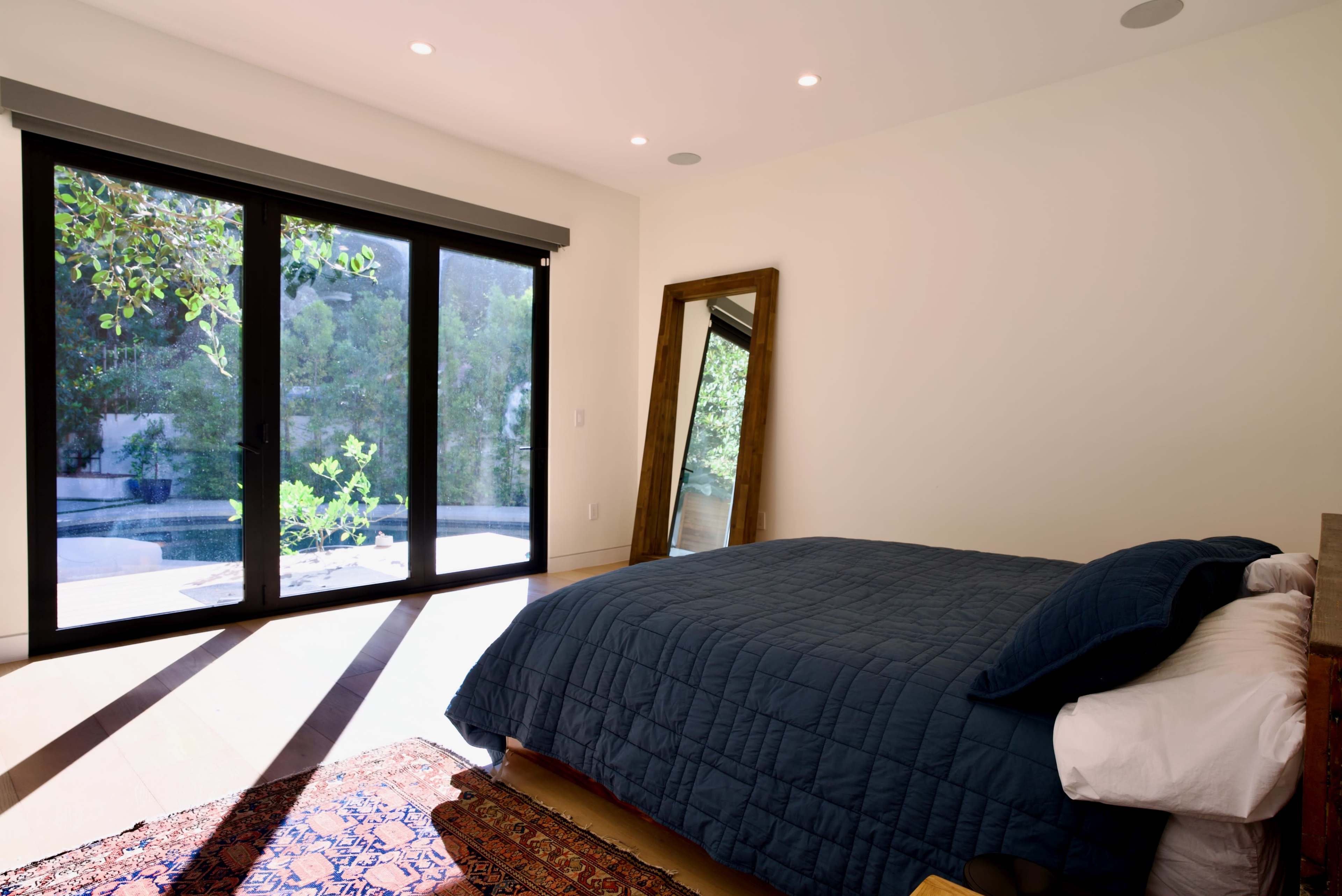 A brightly lit bedroom features a large bed with a navy blue quilt and a full-length mirror beside sliding glass doors that open to a garden view.