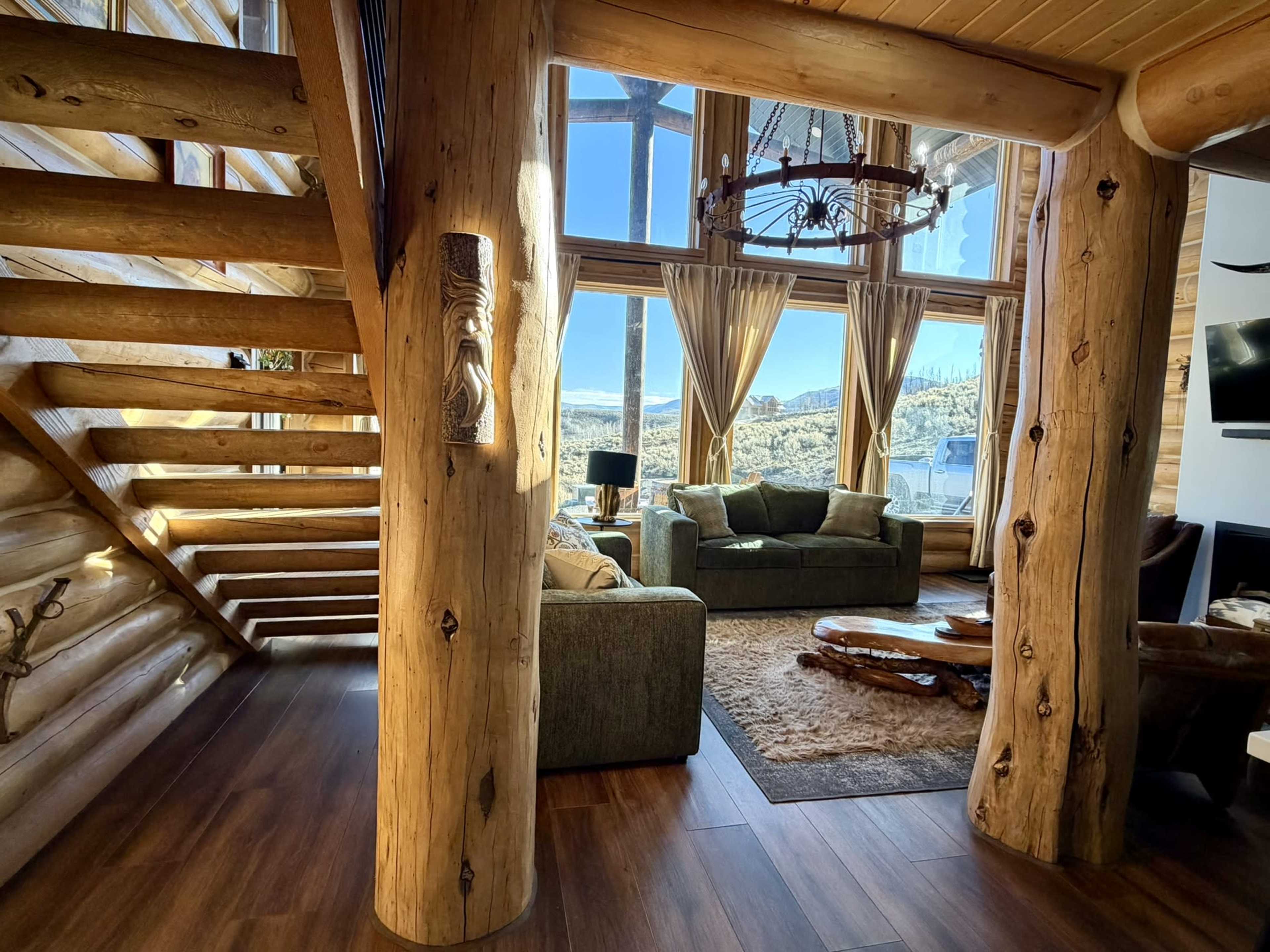 The image shows the interior of a log cabin with a staircase on the left, large windows allowing natural light, and a living area featuring a couch and a wooden coffee table.