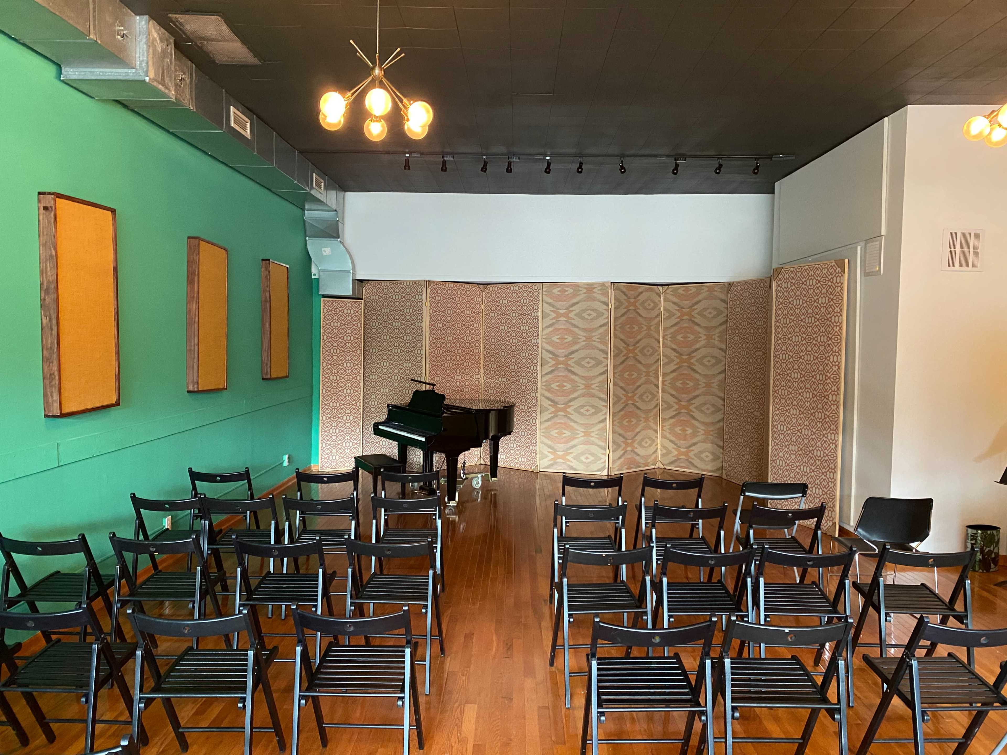 The image shows a small performance space with rows of black chairs facing a grand piano against a green wall, flanked by patterned screens.