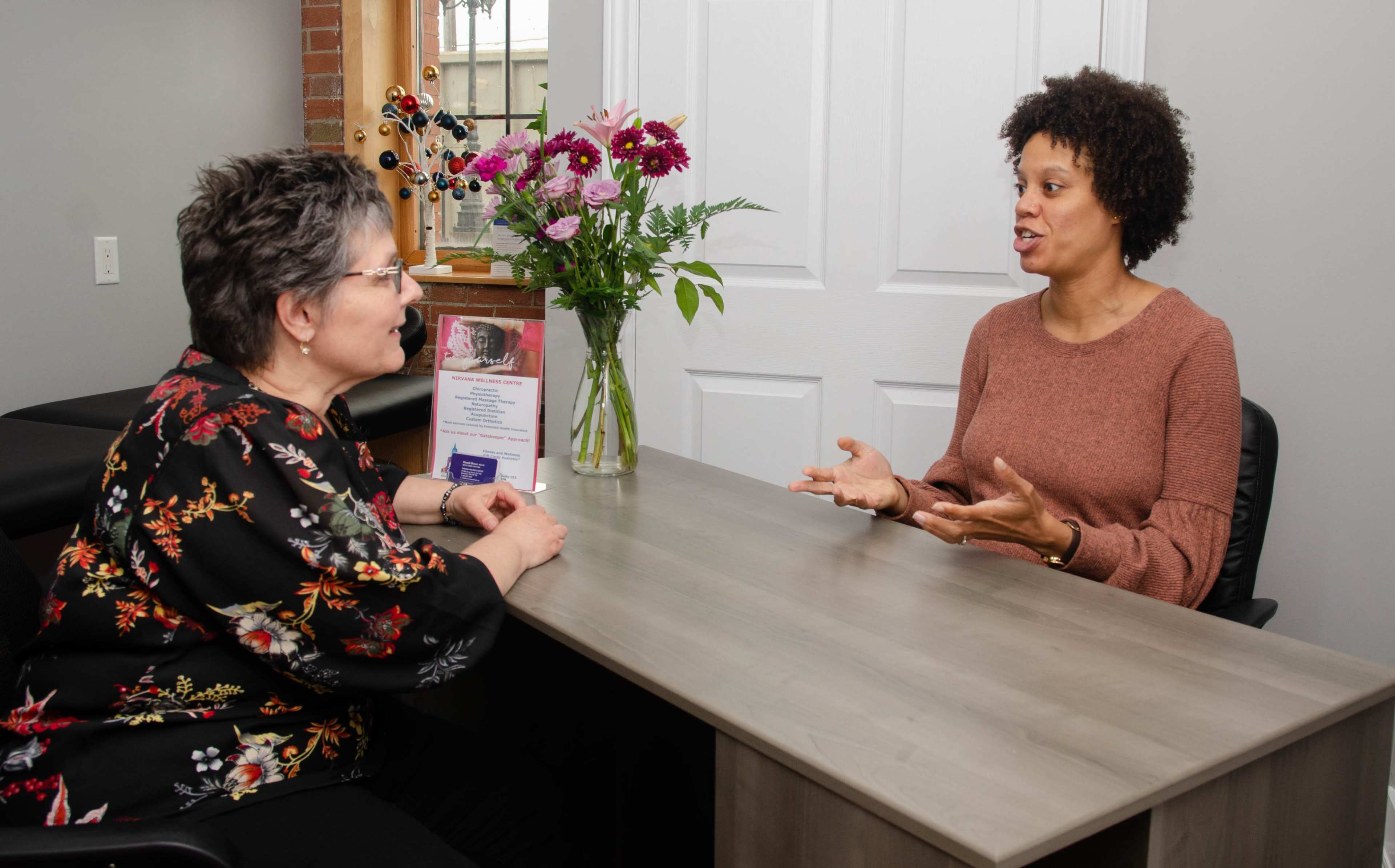 A woman with curly hair speaks to another woman seated at a desk with flowers and promotional materials in the background.
