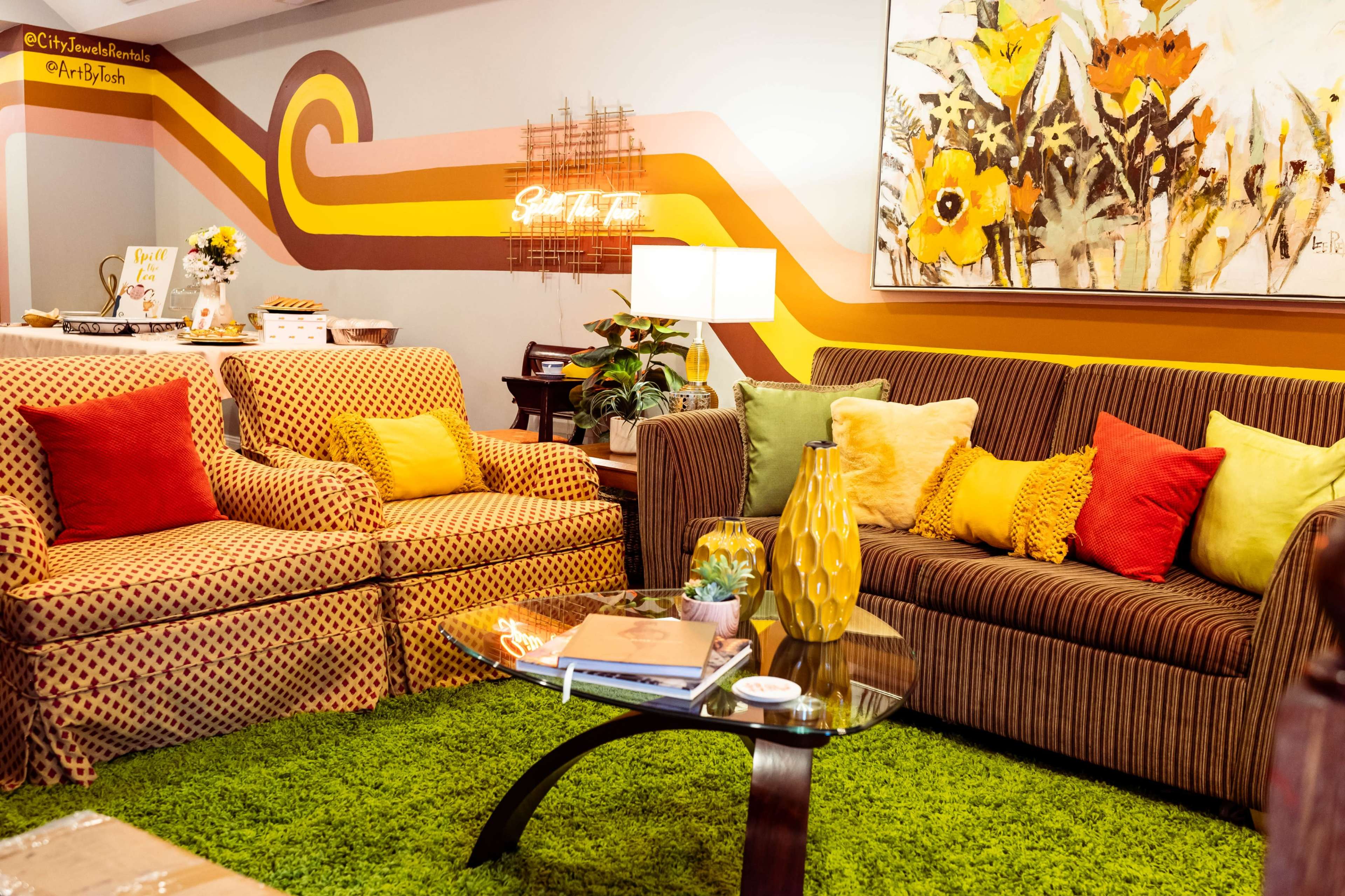 A living room with two sofas, a glass coffee table, and a vibrant, green carpet, set against a decorated wall with colorful patterns and a floral artwork.