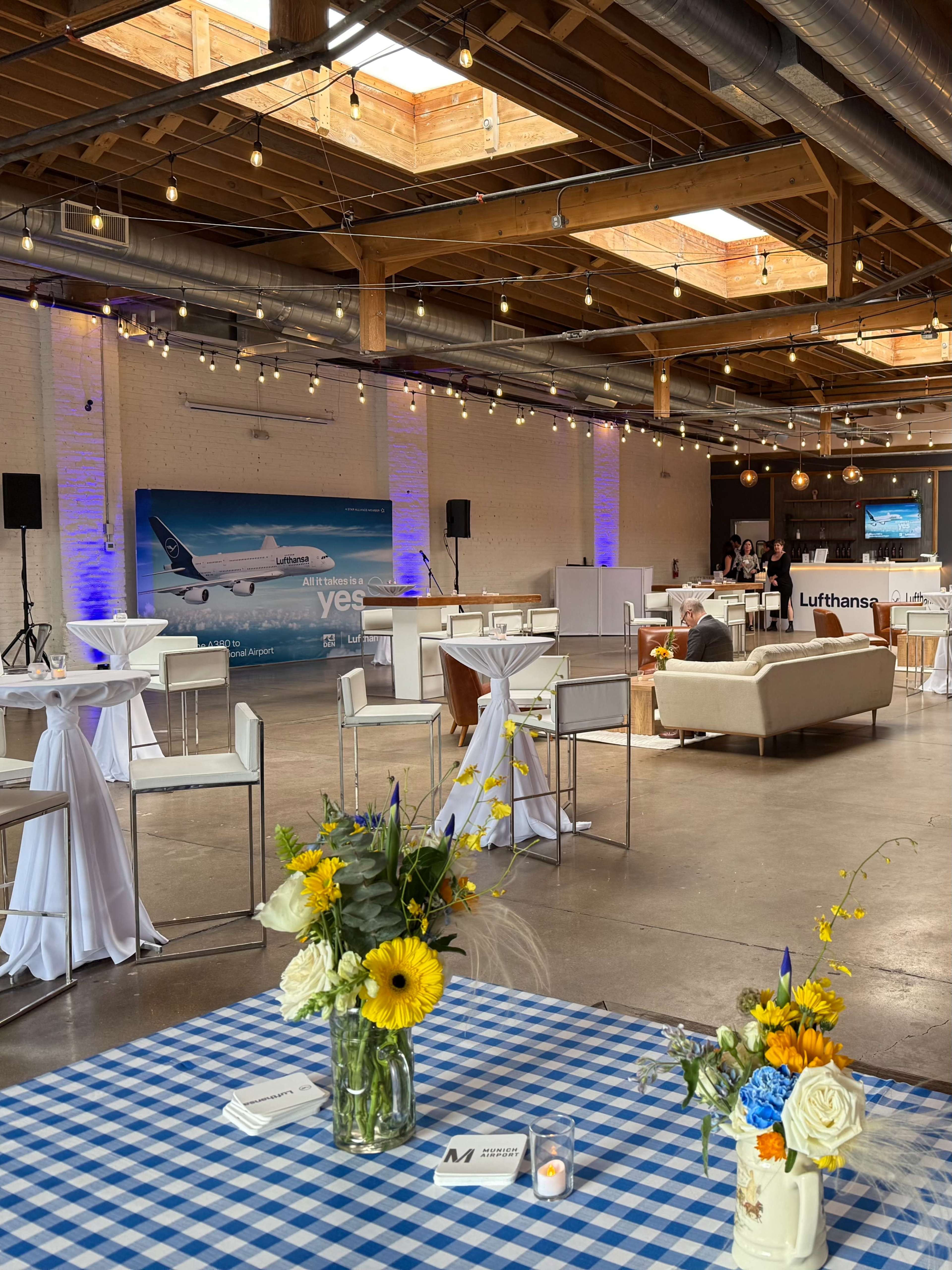 The image depicts a large event space with tables covered in checkered cloth, decorated with floral arrangements, and soft lighting from overhead fixtures.